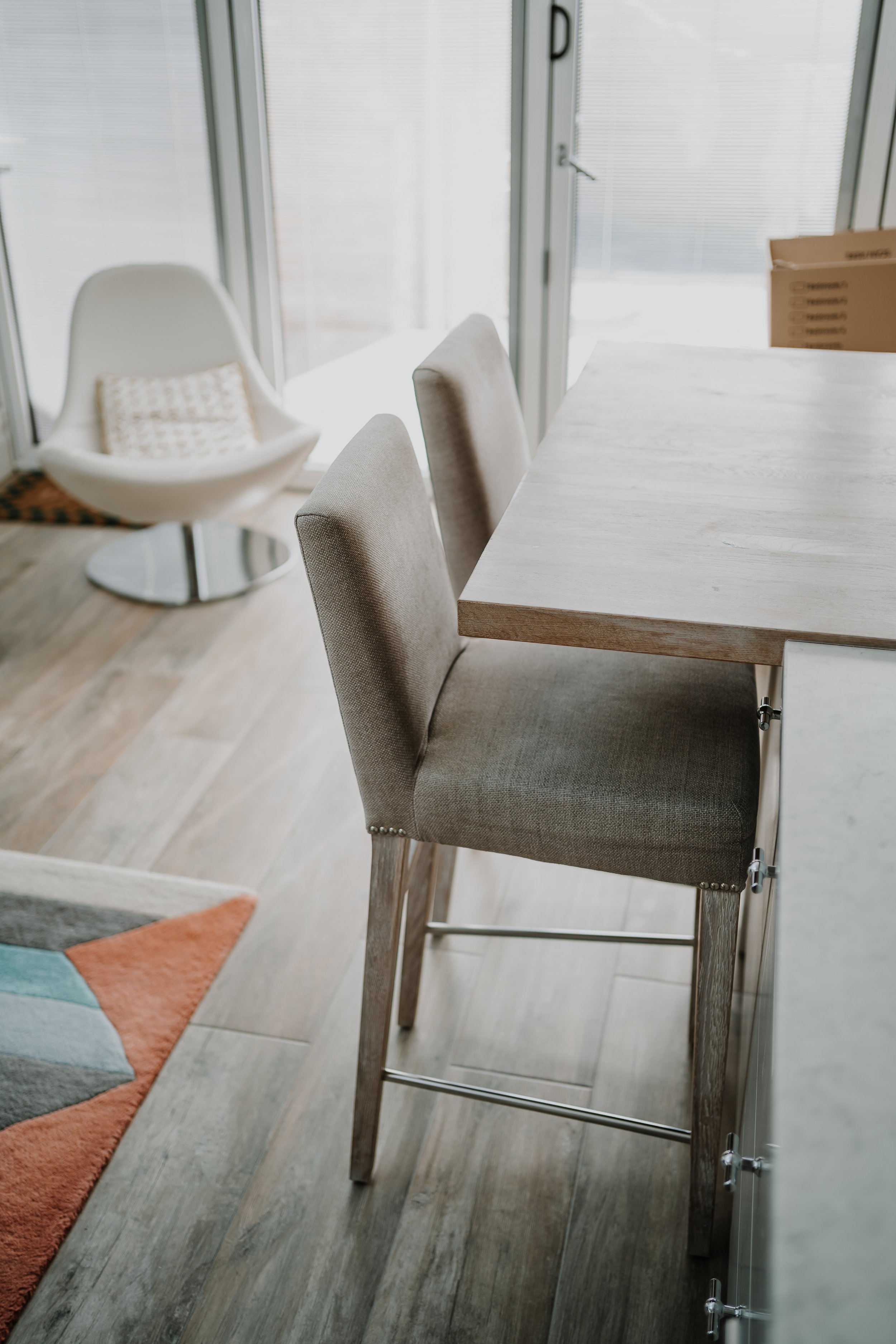 Modern dining area featuring a high-backed upholstered chair at a wooden table, with a white lounge chair and a colorful geometric rug in the background.