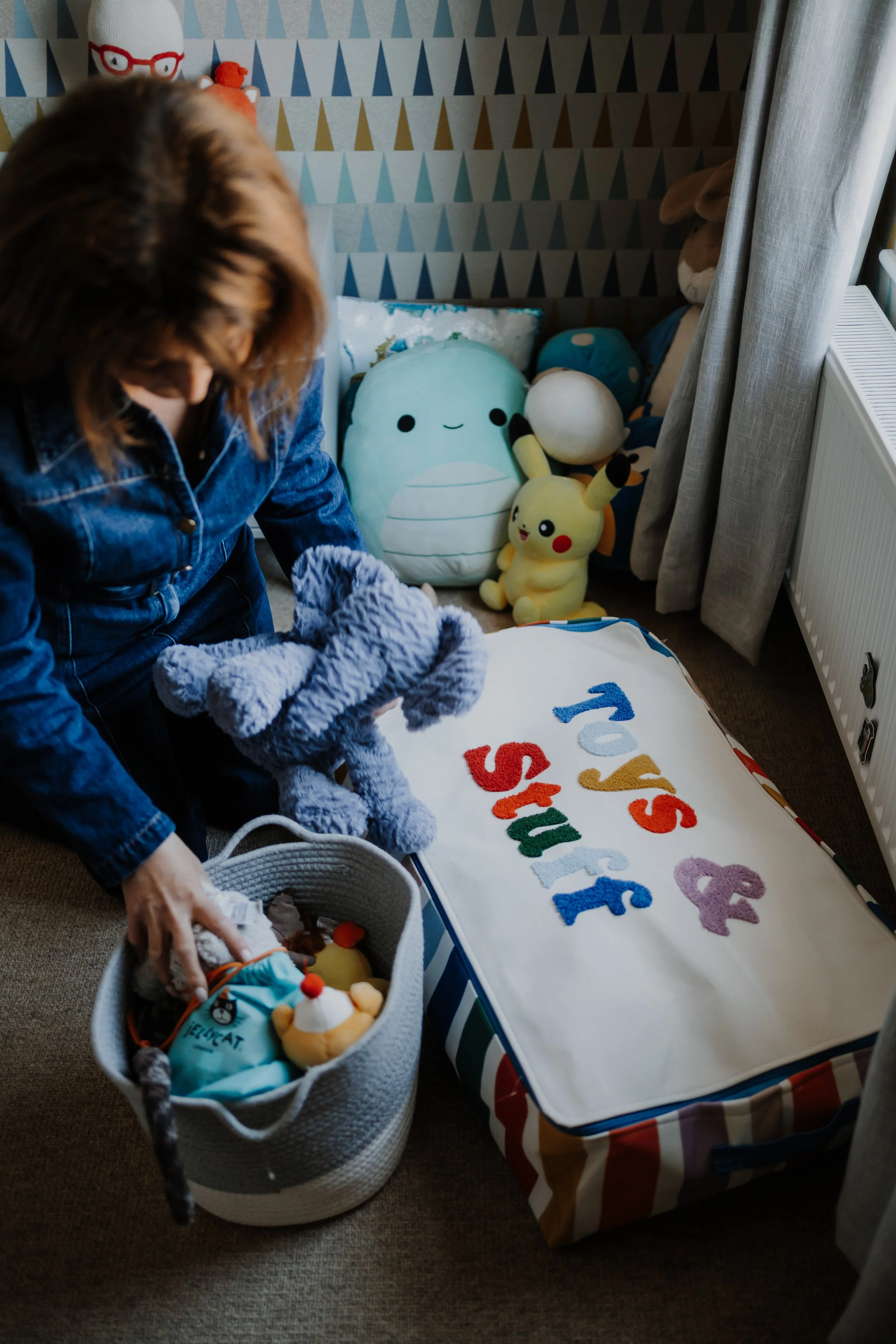 A person packing plush toys and stuffed animals, including Pikachu and a blue whale, into a gray basket. There is a T-Shirt with colorful text on the bed, and a wall with geometric patterned wallpaper in the background.