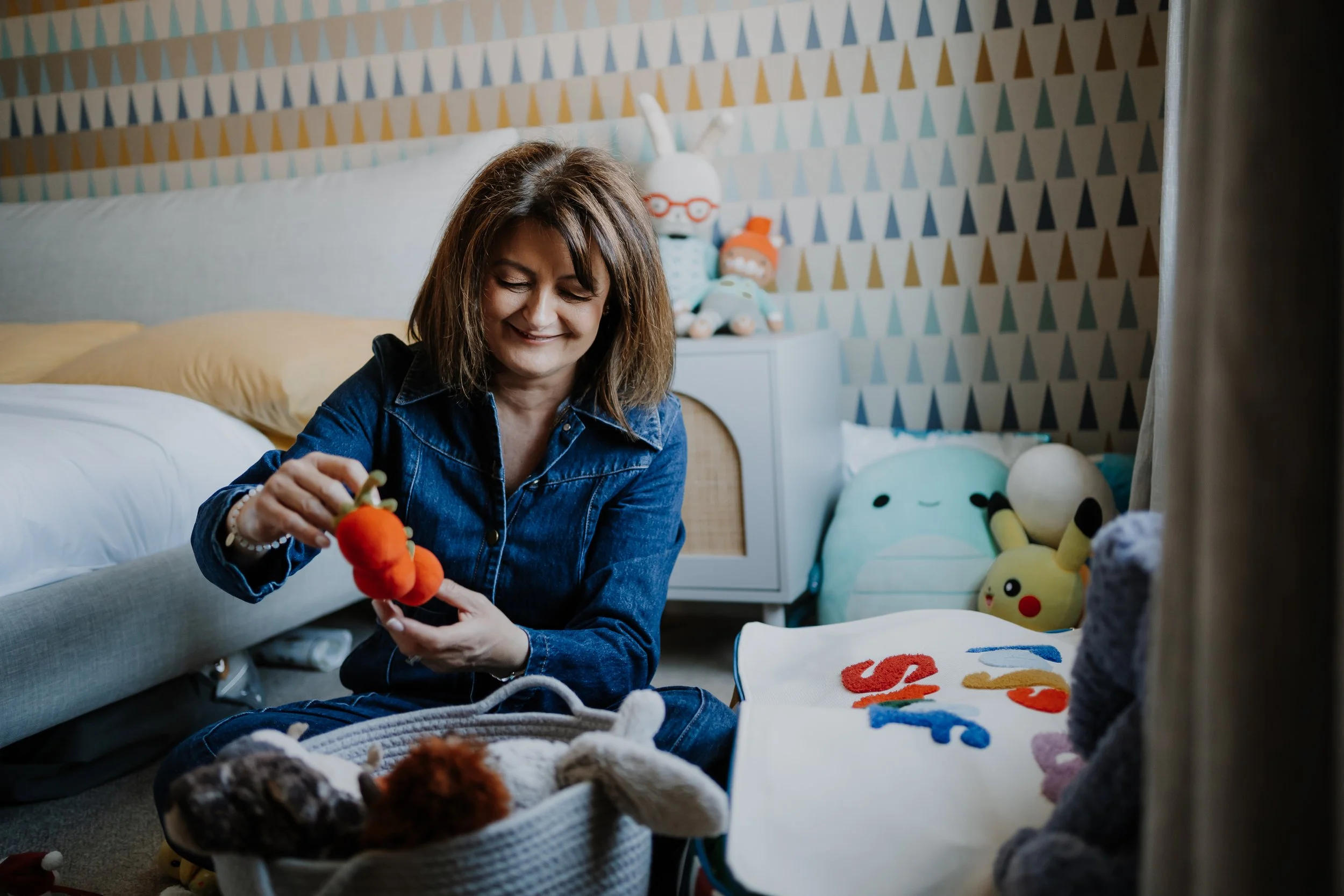 A woman with shoulder-length brown hair, wearing a denim shirt, smiling while holding a plush orange turtle toy in a room decorated with stuffed animals, toys, and colorful decor.