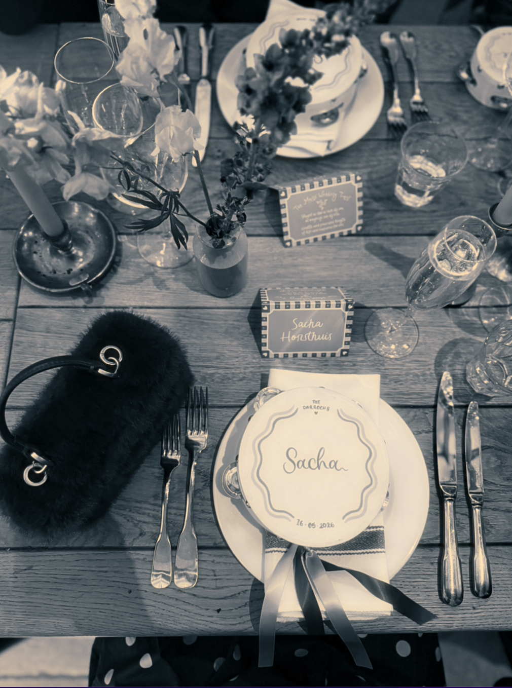 Decorated outdoor dining table set for a celebration, featuring a bouquet of flowers, candles, wine glasses, plates with personalized name cards, and elegant cutlery. The main place setting has a card with the name 'Sacha' and the date '16-05-2016'.