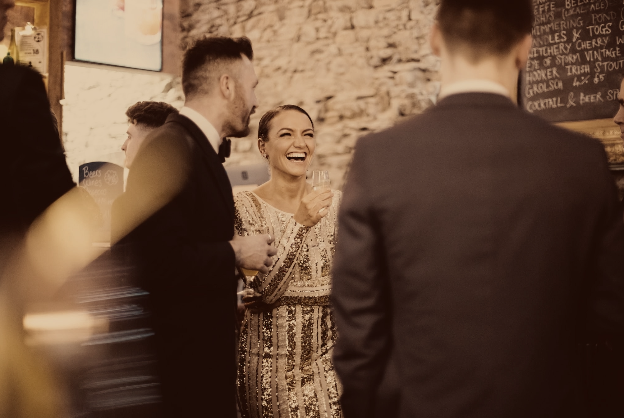 People socializing in a bar or restaurant, with a woman smiling and holding a drink, surrounded by men in formal attire.