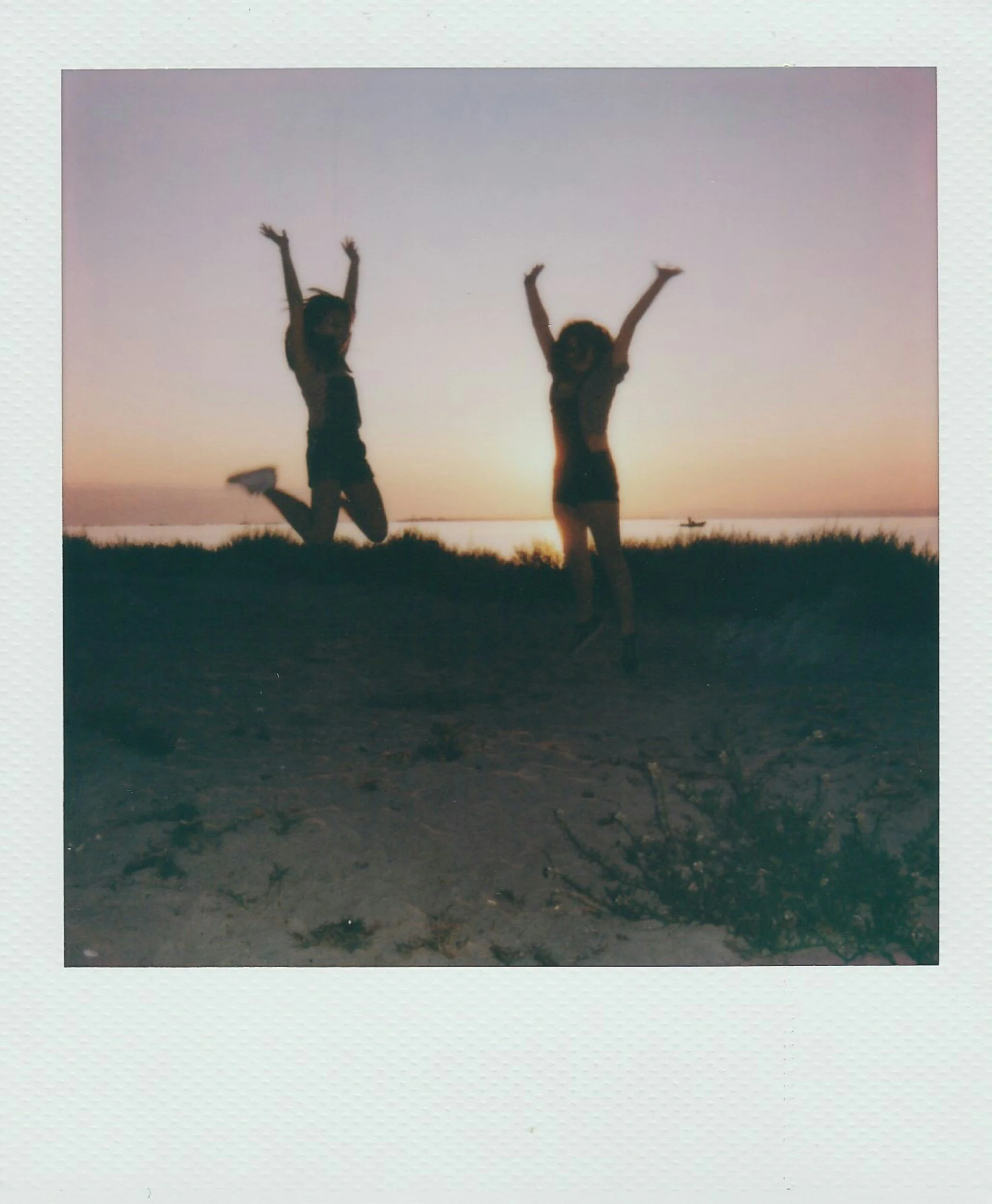 Two children are playing and dancing on a beach at sunset, with their arms raised in the air, in front of a body of water with a boat in the distance.