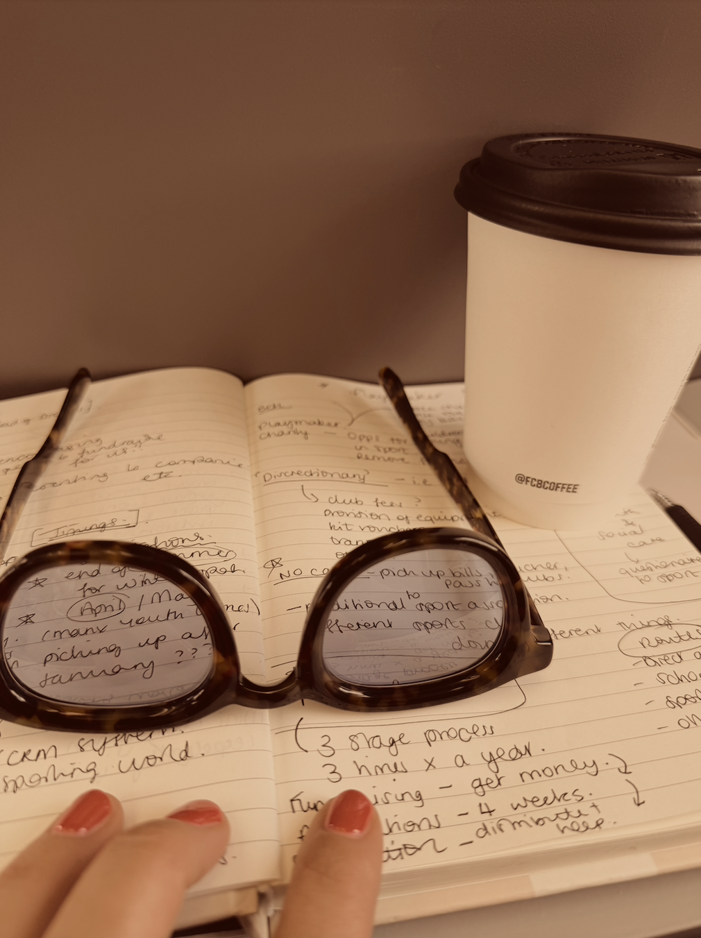 Open notebook with handwritten notes, a pair of glasses resting on it, a paper coffee cup with a brown lid, and a person's hand with painted nails. The note includes sections on 'dictionary' and '3 stage process'.
