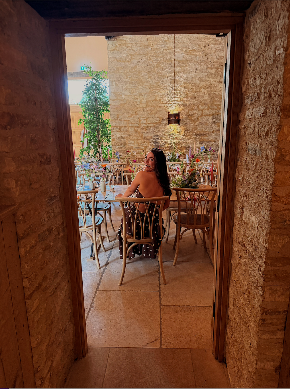 A woman in a polka dot dress sitting at a wooden table in a bright, decorated restaurant with stone walls and blooming flowers.