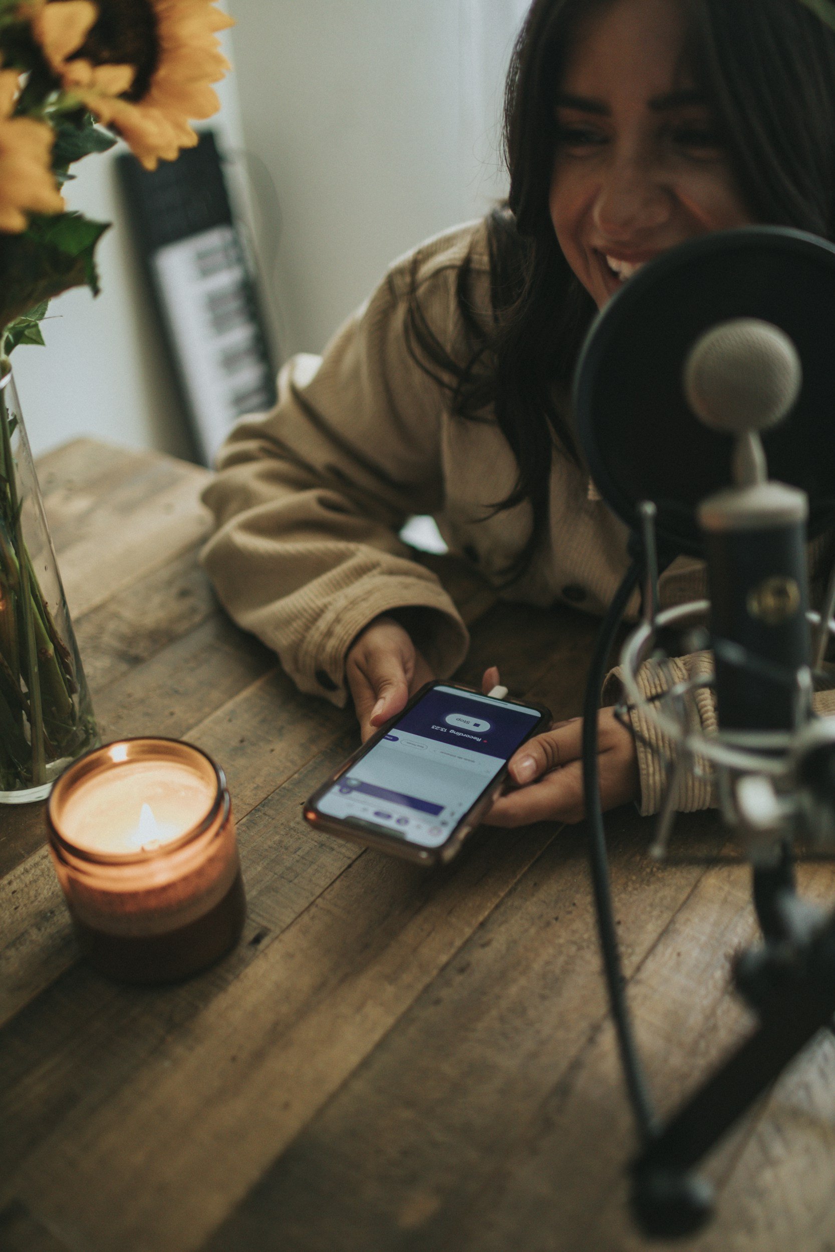 A woman with dark hair smiling and wearing a beige sweater sits at a wooden table with a lit candle, a vase with orange flowers, a candle, a smartphone, and a microphone nearby.
