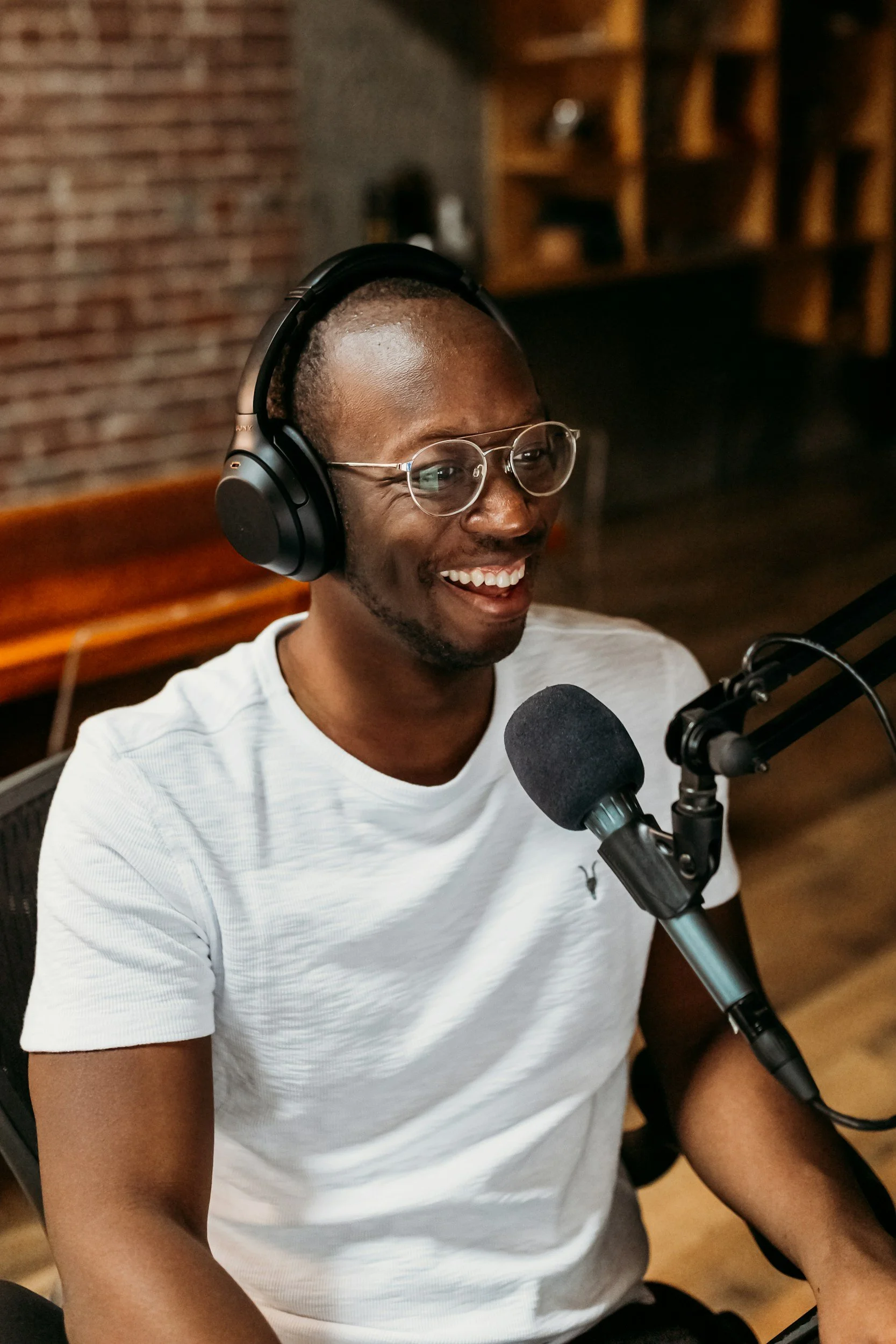 A man wearing glasses and a white t-shirt, smiling while speaking into a microphone, with large headphones on his head, seated in a room with exposed brick wall and wooden shelves in the background.