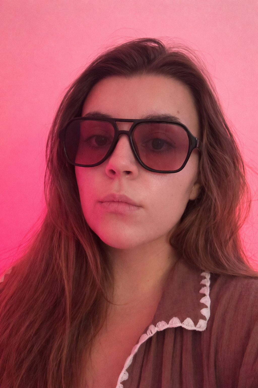 Young woman with long wavy brown hair wearing dark sunglasses and a brown top with white scalloped collar against a pink background.
