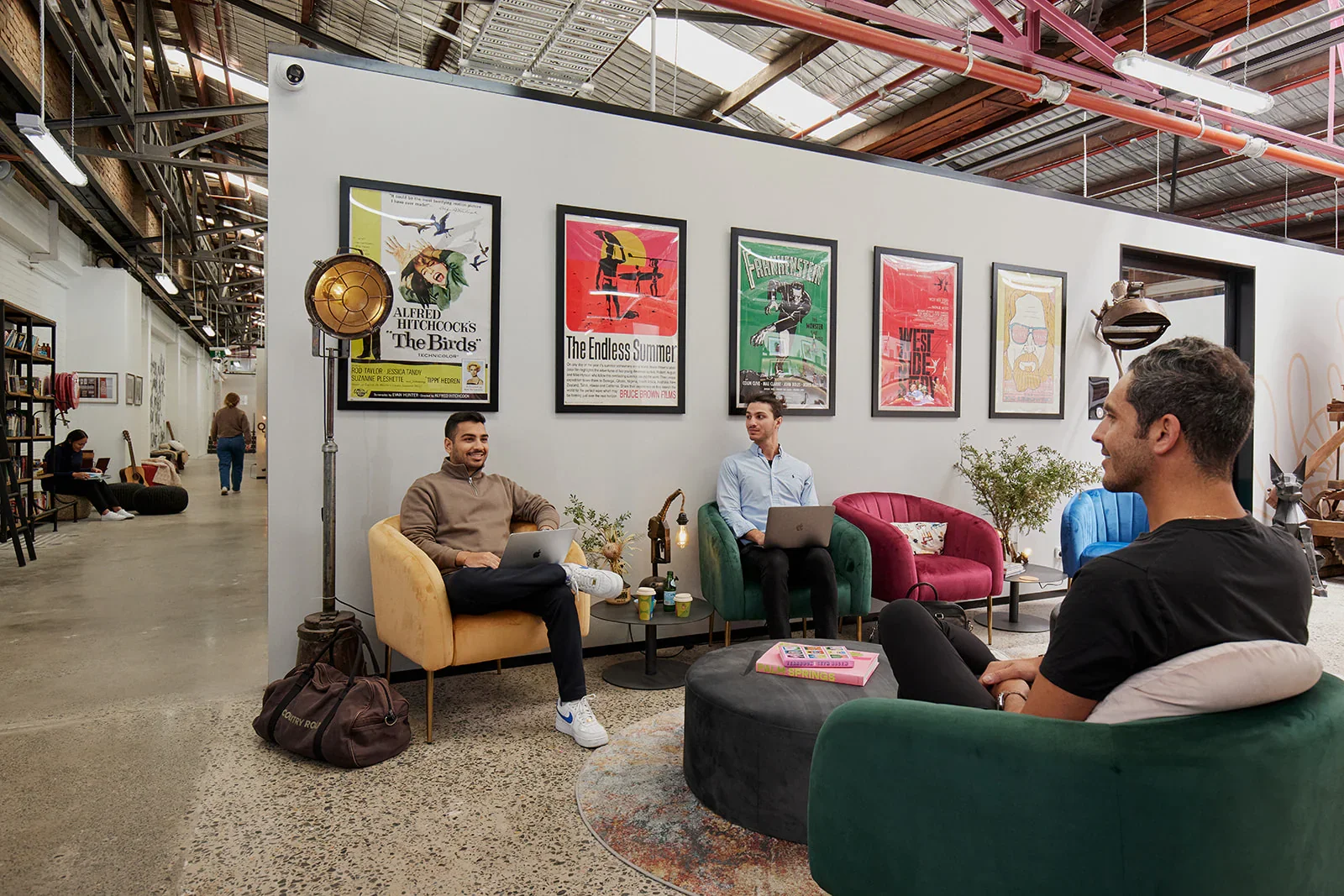 Three men sitting in a casual lounge area, two with laptops, one with a notebook, engaging in conversation. The space is decorated with colorful chairs, potted plants, framed posters on the wall, and a vintage-style standing lamp. In the background, people are walking and sitting at tables in an open, industrial-style space.