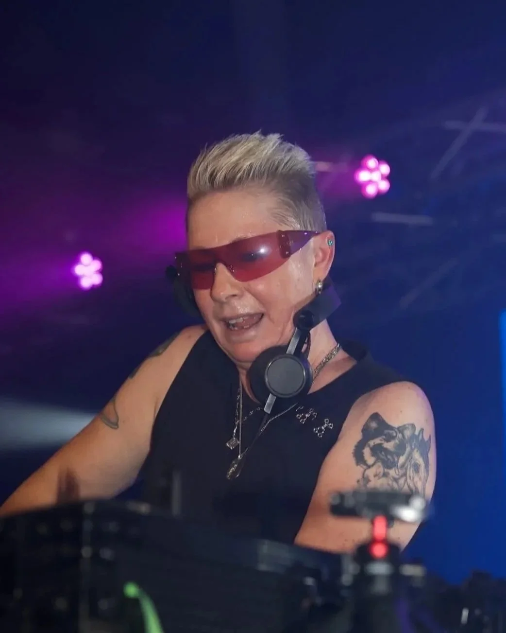 A DJ with short platinum blonde hair, sunglasses, and tattoos on their arms, wearing headphones around their neck, performing at a nightclub with purple and pink stage lights.