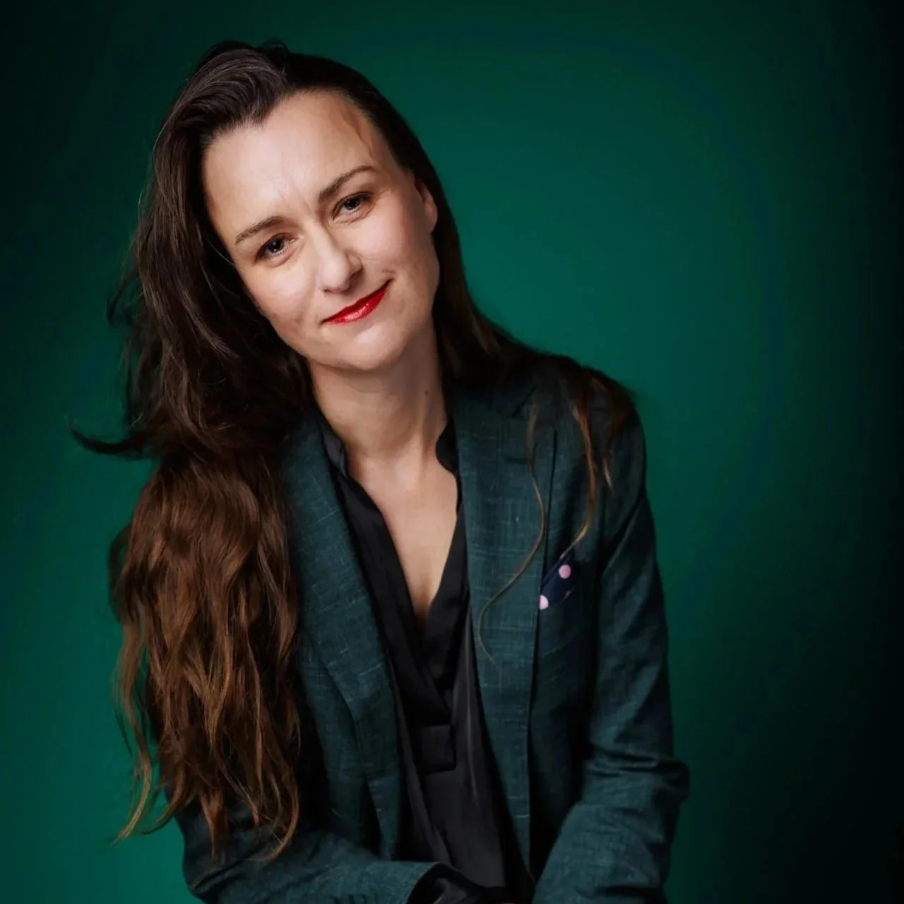 A woman with long brown hair and fair skin wearing a dark blazer and black shirt, smiling, against a green background.