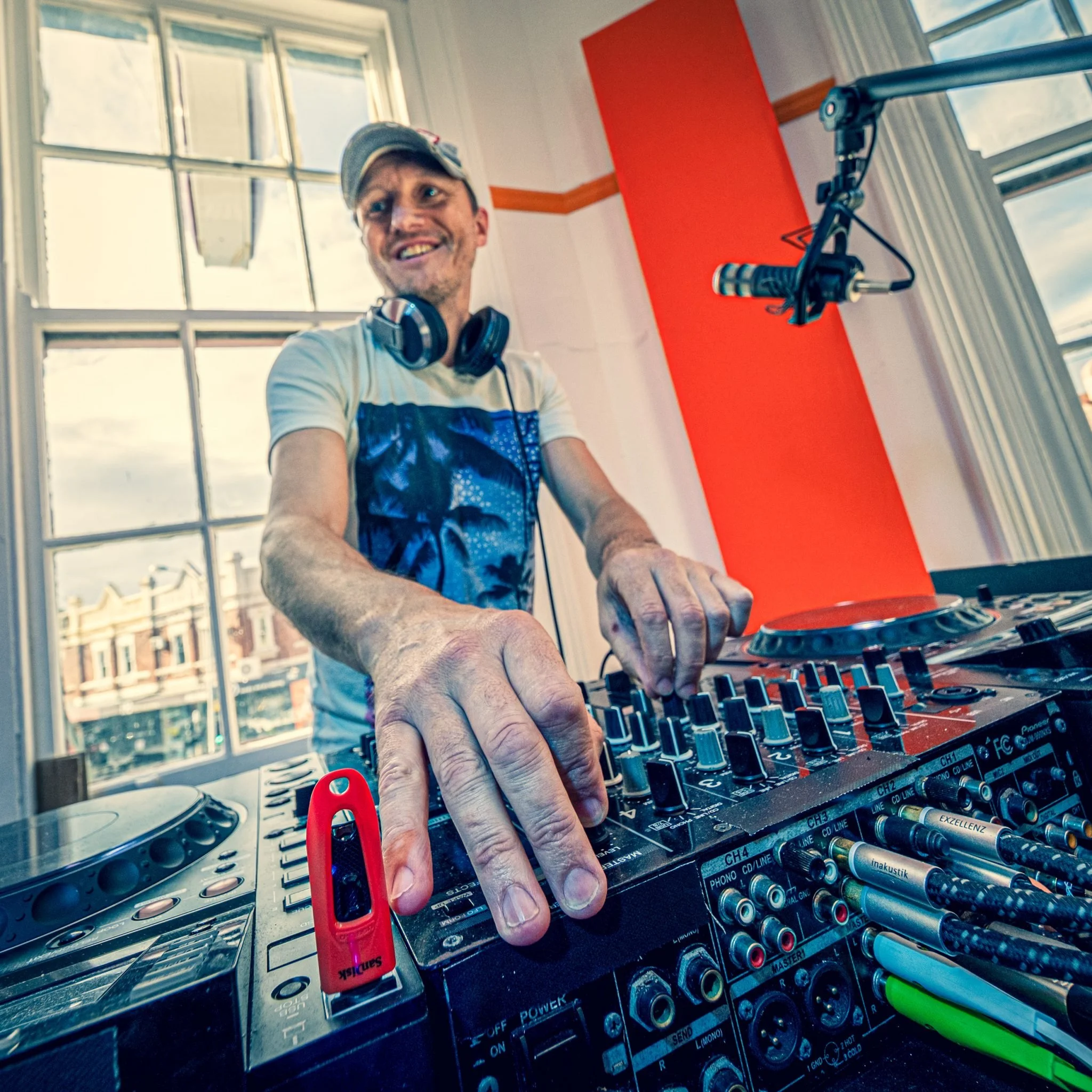 A man wearing a cap and headphones is DJing with a mixer and various colored markers on a table. He is smiling, inside a room with large windows and a bright orange wall.