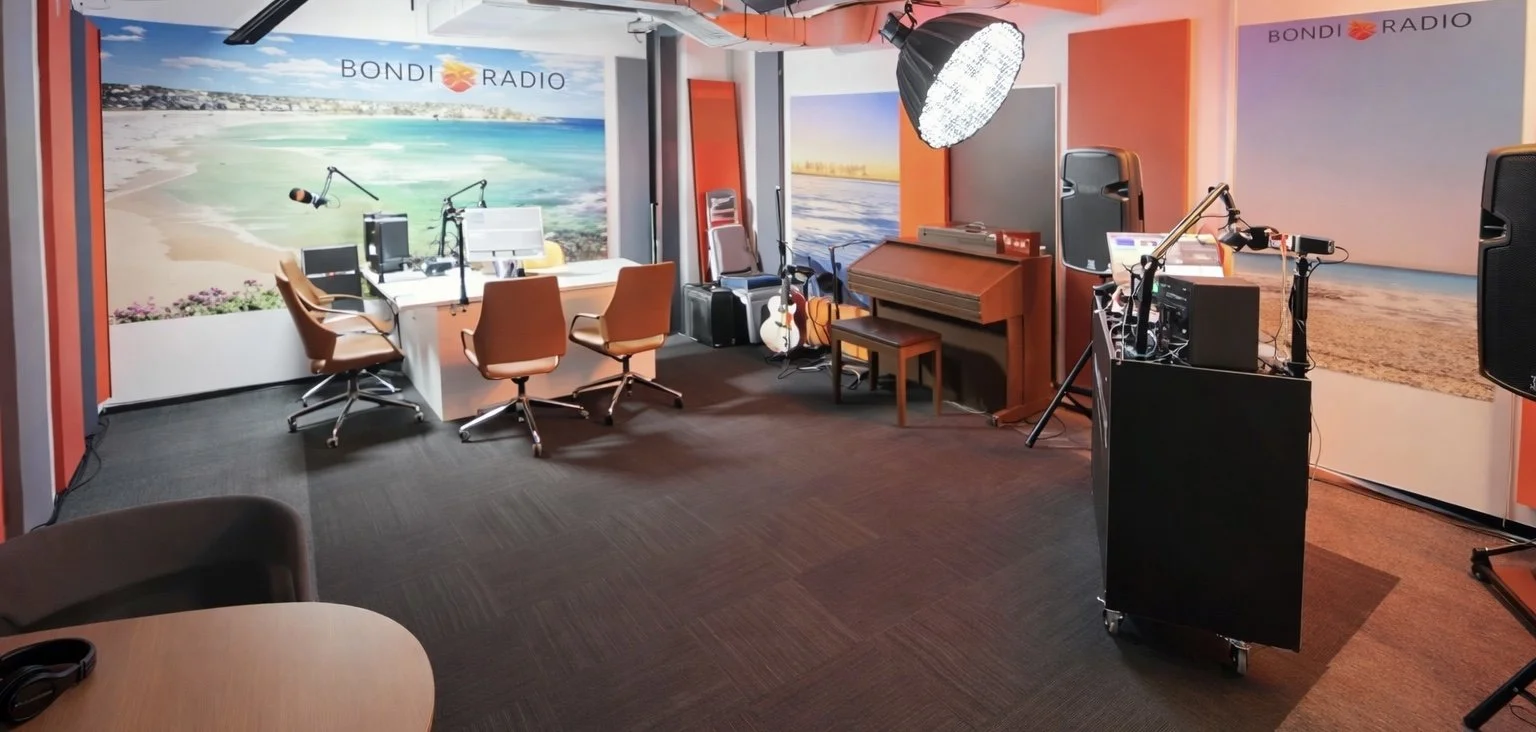 Radio station or recording studio with 'Bondi Radio' branding, microphones, headphones, a piano, guitars, and large beach-themed murals on the walls.
