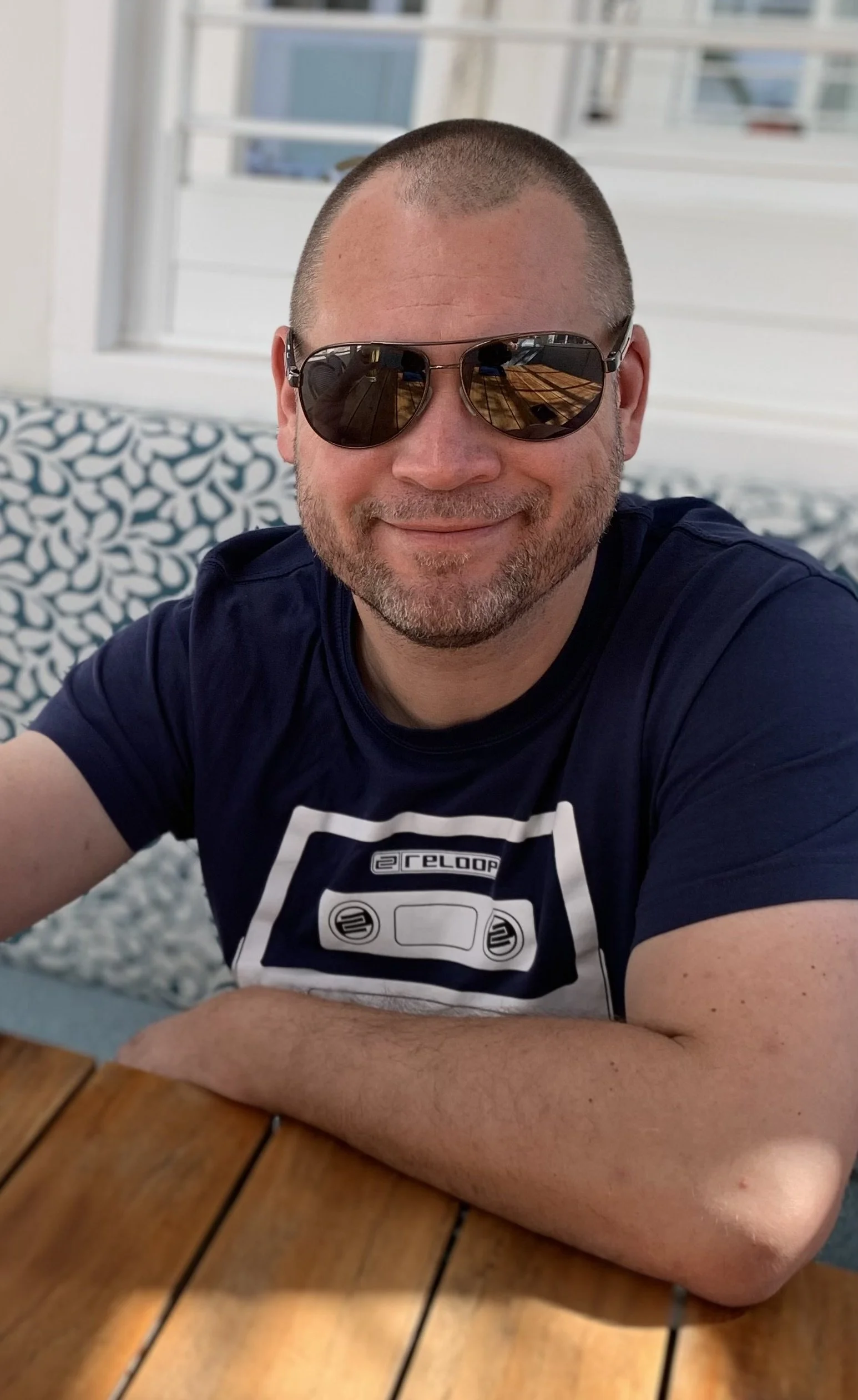 A man with a shaved head and beard wearing sunglasses, smiling, sitting at a wooden table with a patterned cushion and window with blinds in the background.