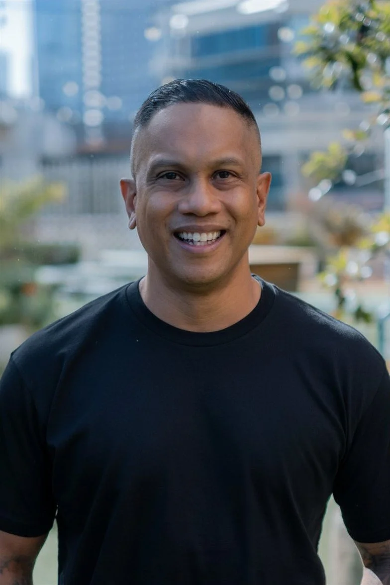 A smiling man with short dark hair in a black t-shirt, standing outdoors with a cityscape background including buildings and trees.