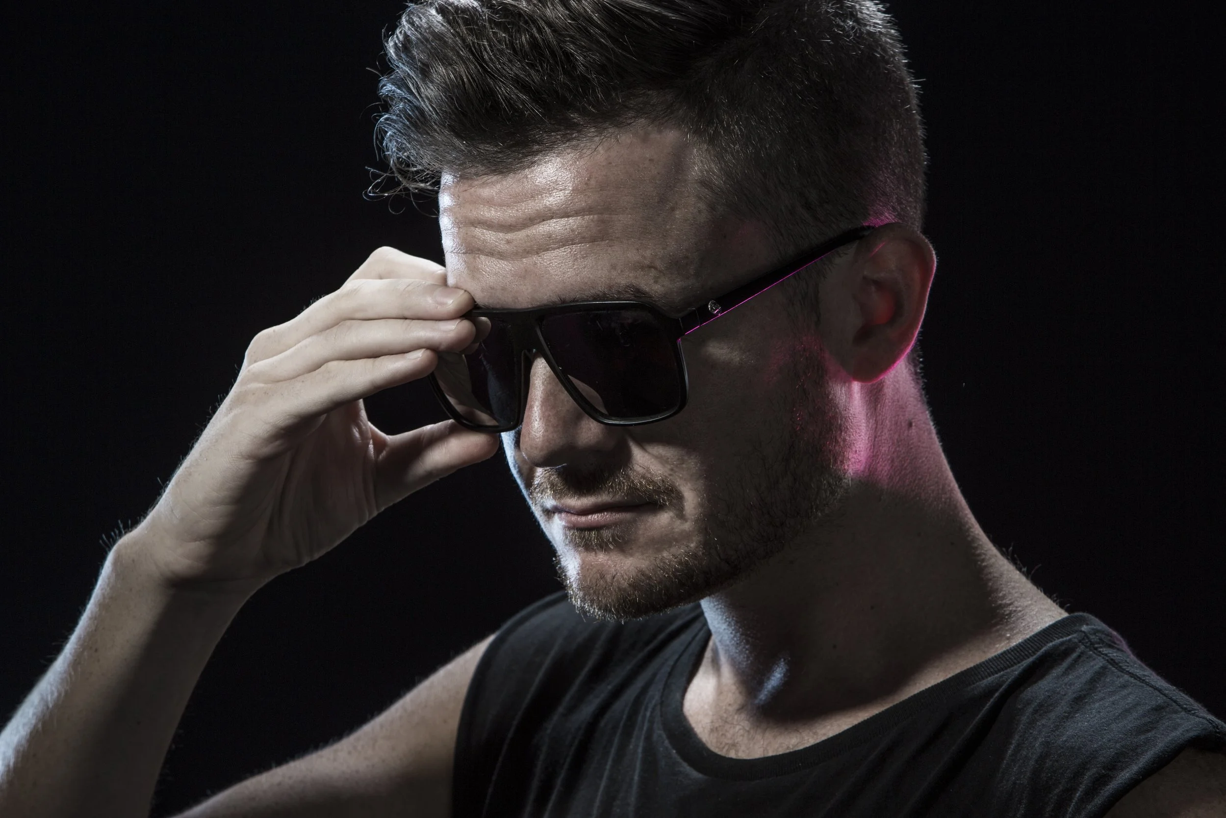 A young man with short dark hair and facial hair, wearing sunglasses and a black sleeveless shirt, adjusting his sunglasses against a dark background.