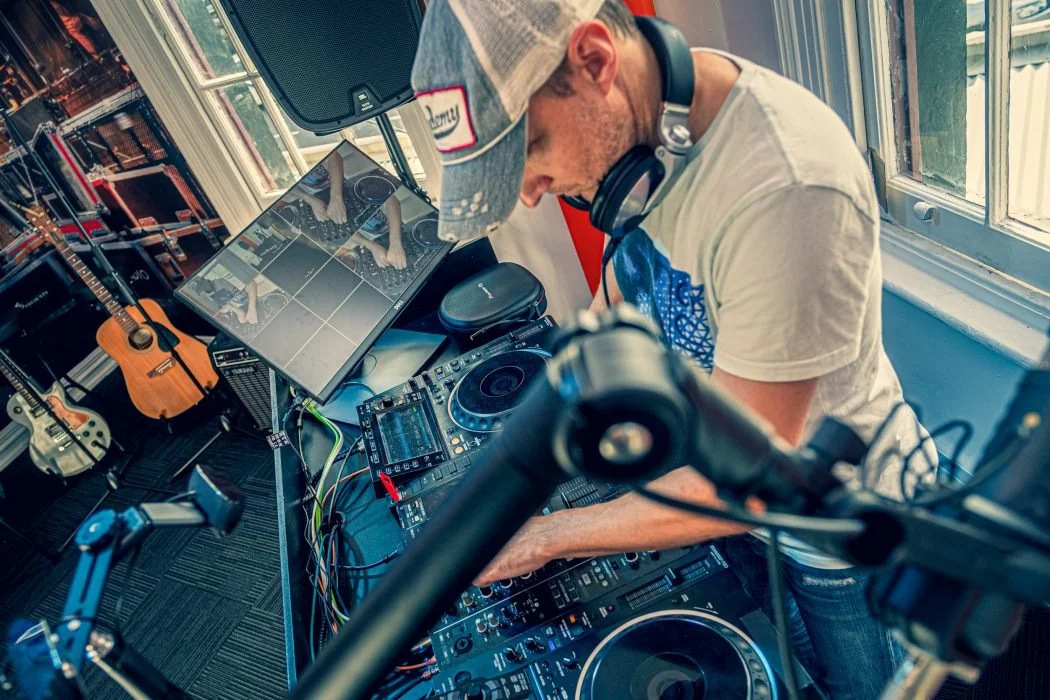 A person wearing a baseball cap and headphones is DJing with a mixer and turntables in a room filled with guitars, a monitor, and large windows.