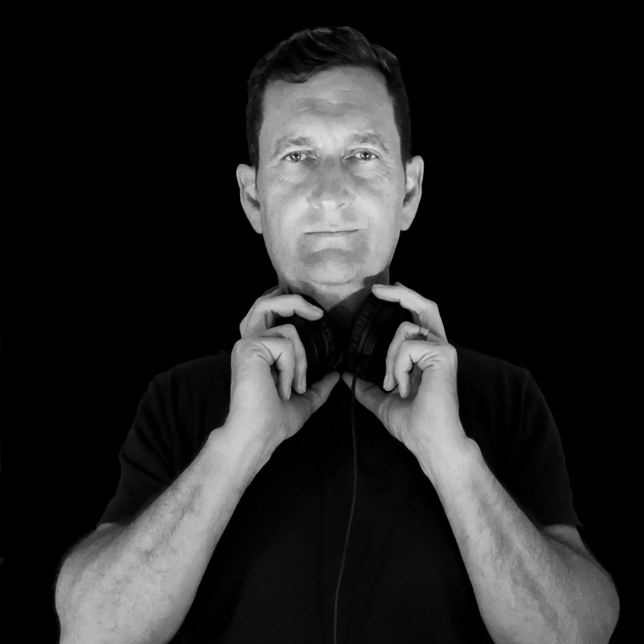 Black and white image of a man with short hair, holding a zipper collar with both hands, wearing a black shirt, looking directly at the camera against a black background.