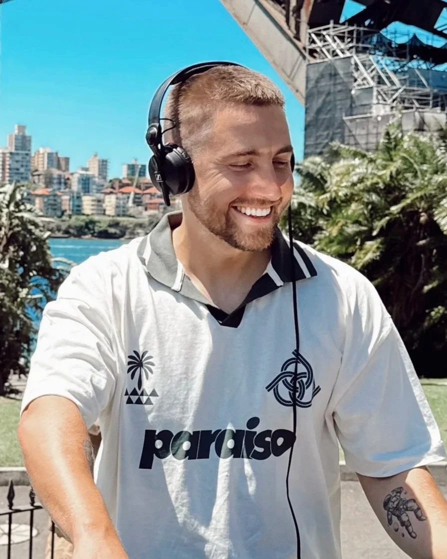A smiling man with a beard and short hair wearing a white sports jersey with logos, listening to headphones outdoors during daytime, with a city skyline and skyscrapers in the background.