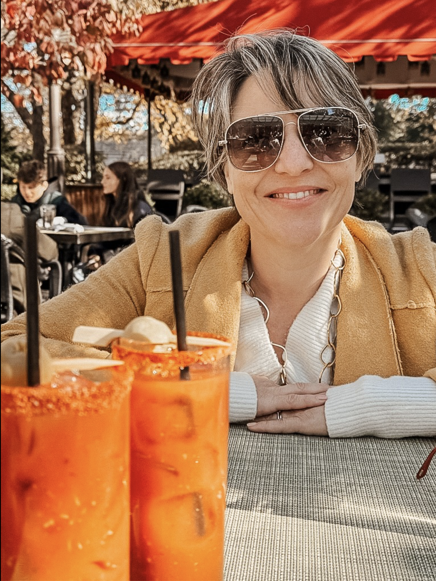 A woman with short gray hair and sunglasses smiling at the camera while sitting at an outdoor restaurant or cafe. There are two glasses of orange-colored drinks with black straws in front of her, and other people are seated at tables in the background under red umbrellas, with trees and blushing flowers nearby.