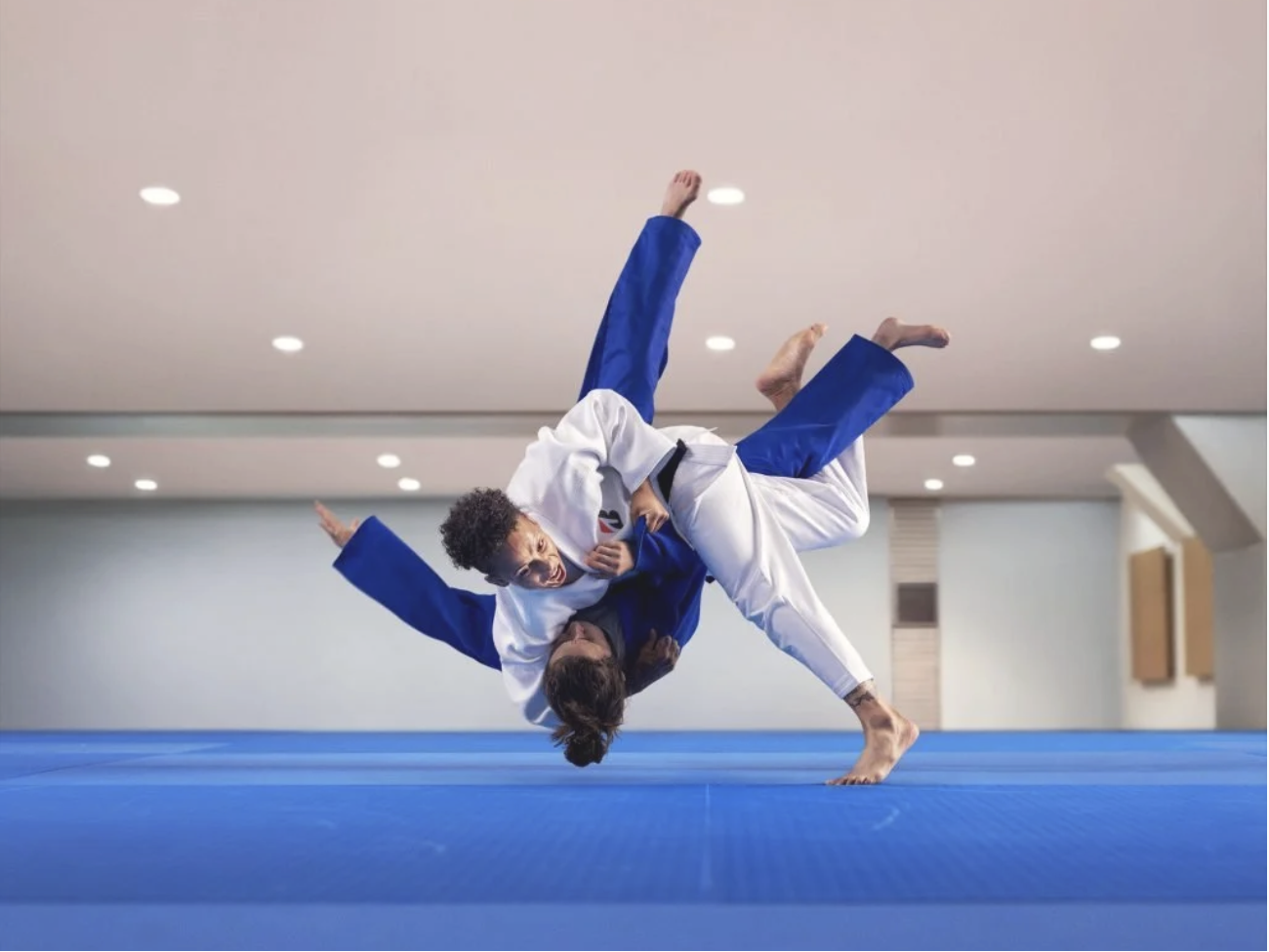 Two individuals practicing Brazilian Jiu-Jitsu on a blue mat, engaged in a grappling move, inside a gym or dojo with ceiling lights.