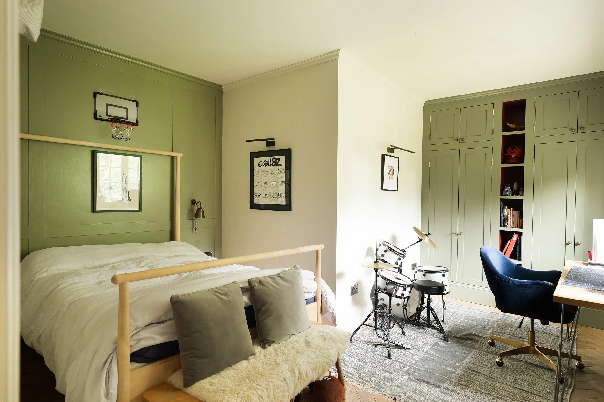 A bedroom with green walls, a bed with white bedding and pillows, framed artwork on the wall, a drum set with a stool, a blue chair, a wooden desk, and built-in green cabinets with books and decor.