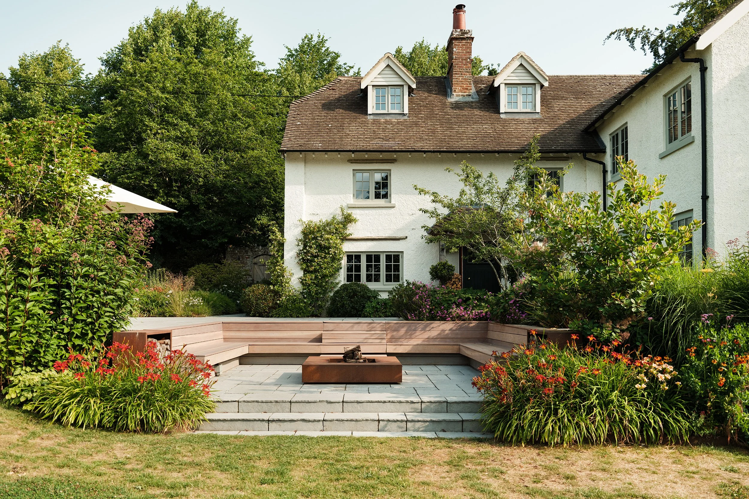A backyard with a stone patio, a built-in bench, a firepit, and well-maintained flower beds with colorful flowers. There are steps leading up to the backyard, and a white house with a gabled roof and multiple windows is seen in the background, surrounded by trees and greenery.