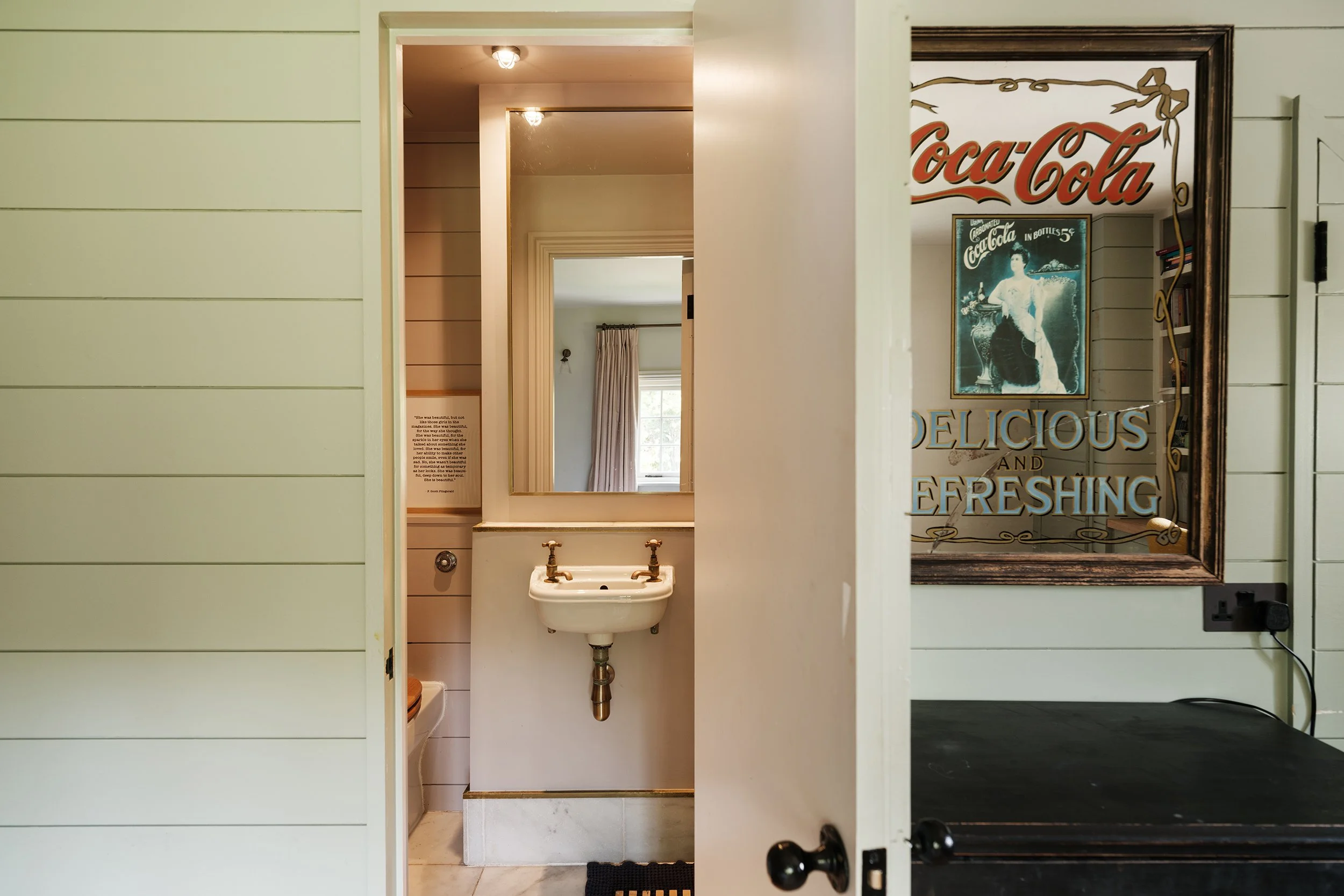 A view of a small bathroom with a white sink and mirror, visible through an open doorway. The bathroom has cream-colored walls with horizontal paneling. On the right side of the image, a large vintage Coca-Cola sign is reflected in a mirror, and a window with curtains is visible in the background.