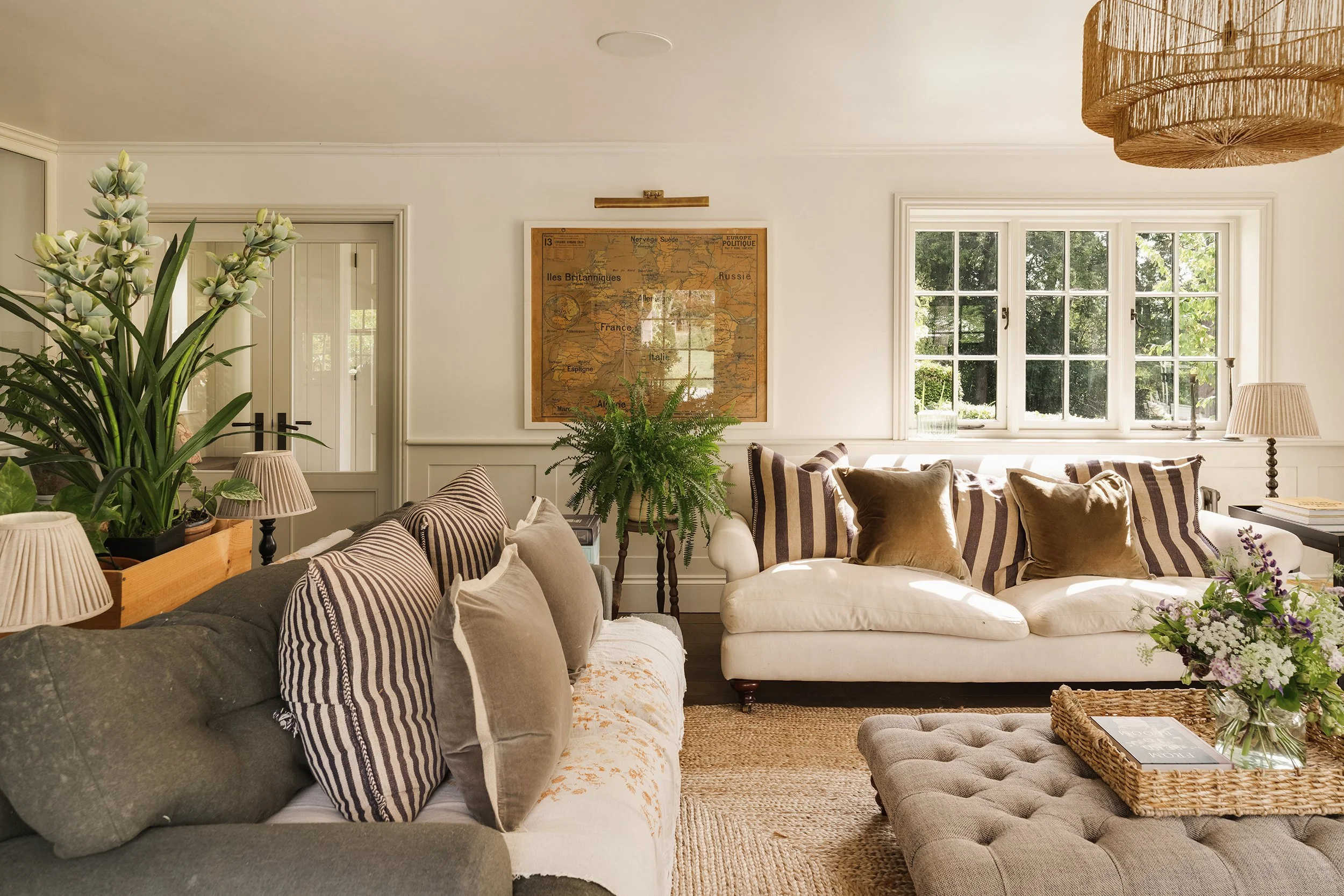 Living room with white walls, large window with greenery outside, framed map on the wall, and a variety of sofas and chairs with cushions. There are plants, lamps, and a woven basket on a table.