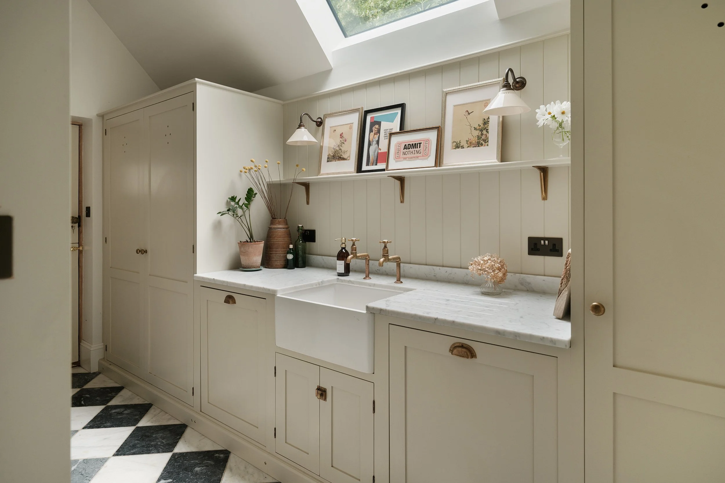 A cozy kitchen corner with white cabinetry, a marble countertop, and a skylight overhead. Decor includes framed artwork, a small pitcher with flowers, green bottles, and brass plumbing fixtures.