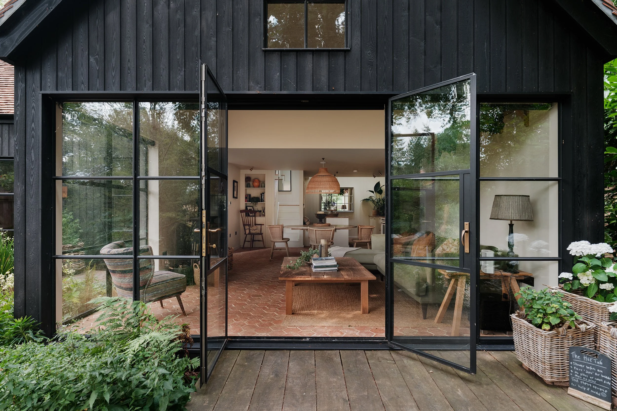 View of an open black-framed glass door leading into a cozy, modern living space with wooden furniture, indoor plants, and decorative items, surrounded by lush greenery.