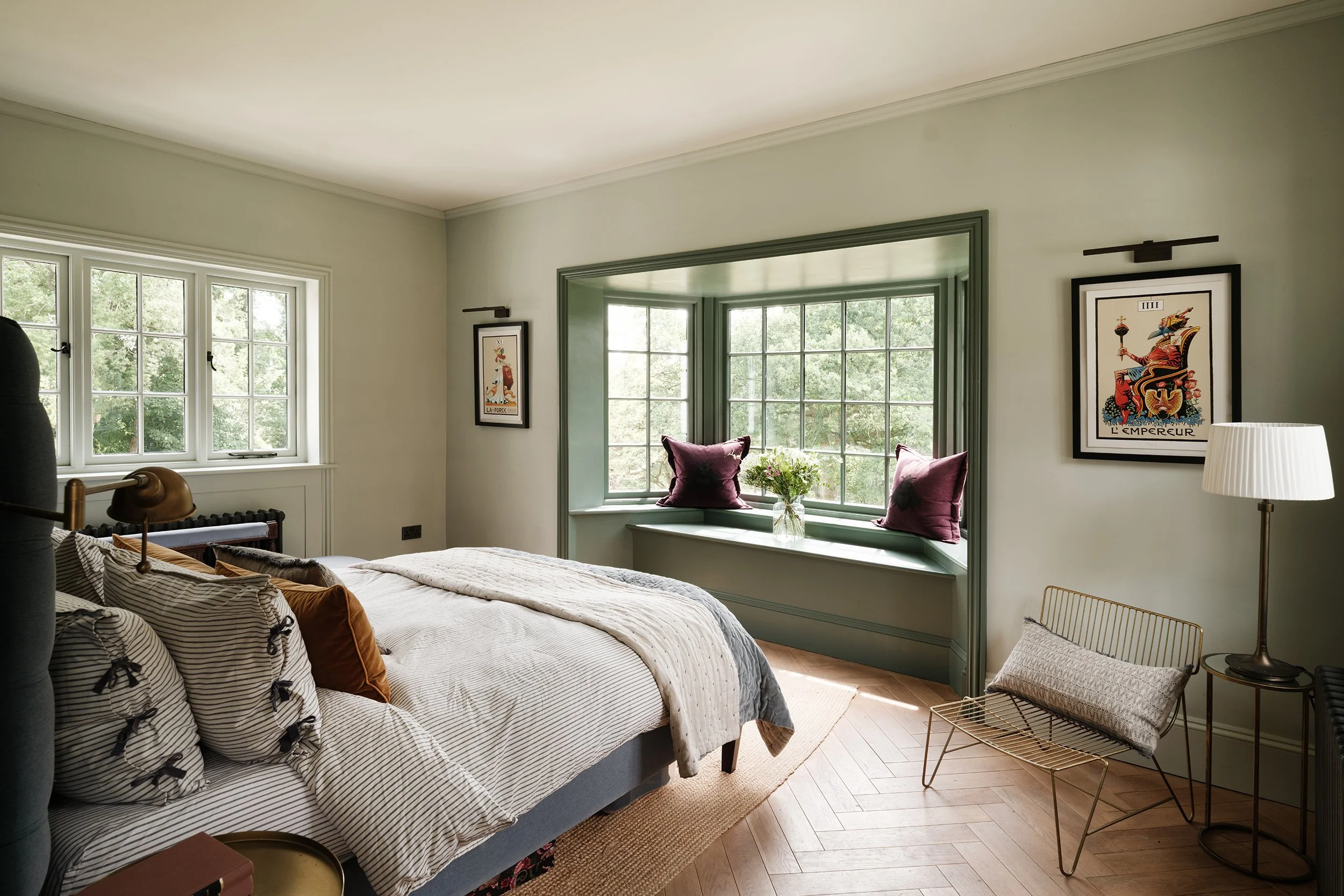 A cozy bedroom with a large bed, striped pillows, and a soft blanket. Sunlight streams through multiple windows, illuminating a built-in window seat with purple cushions and a vase of flowers. The room features framed artwork, a white lamp, and a gold wire chair, with light-colored wooden floors.