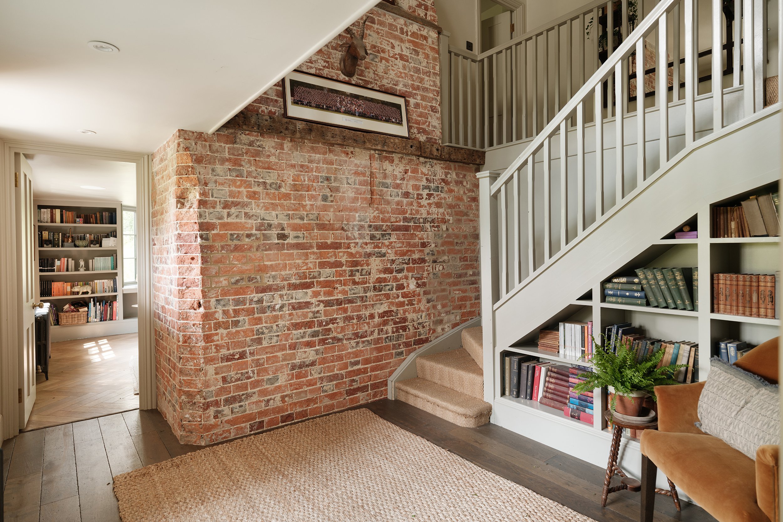 Living room with exposed brick wall, staircase with white railing, built-in white bookshelf filled with books, and a potted fern plant.