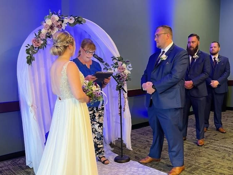 A wedding ceremony with a bride and groom exchanging vows in front of an officiant, under a floral arch, with three groomsmen standing behind.