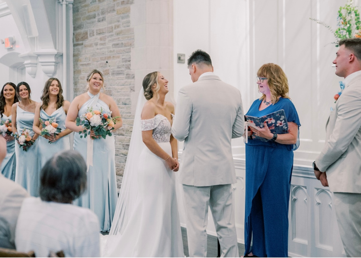 A bride and groom exchange vows during their wedding ceremony, with bridesmaids and groomsmen standing nearby, and an officiant reading from a book.