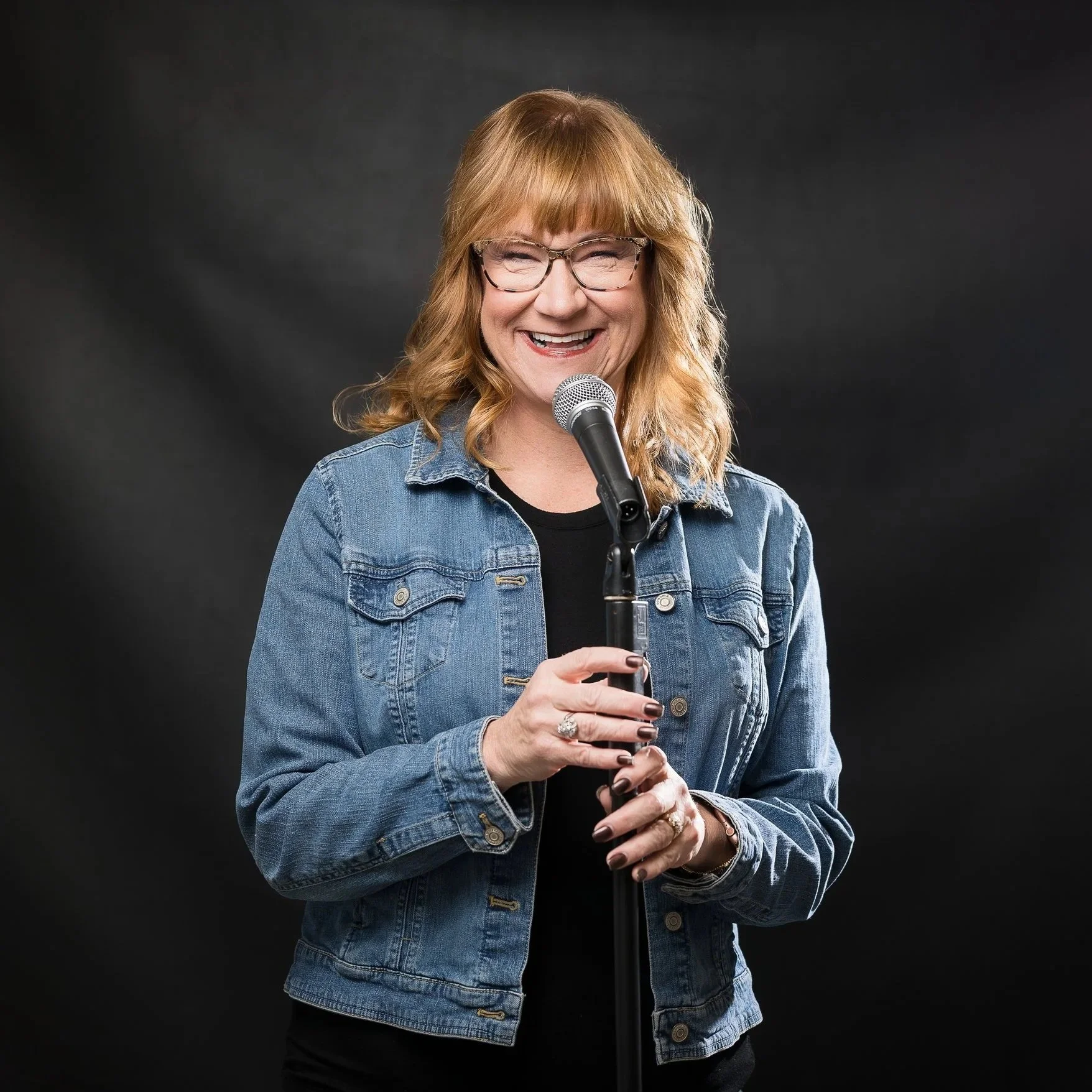 A smiling woman with glasses and orange hair, wearing a denim jacket, standing in front of a black background, holding a microphone.