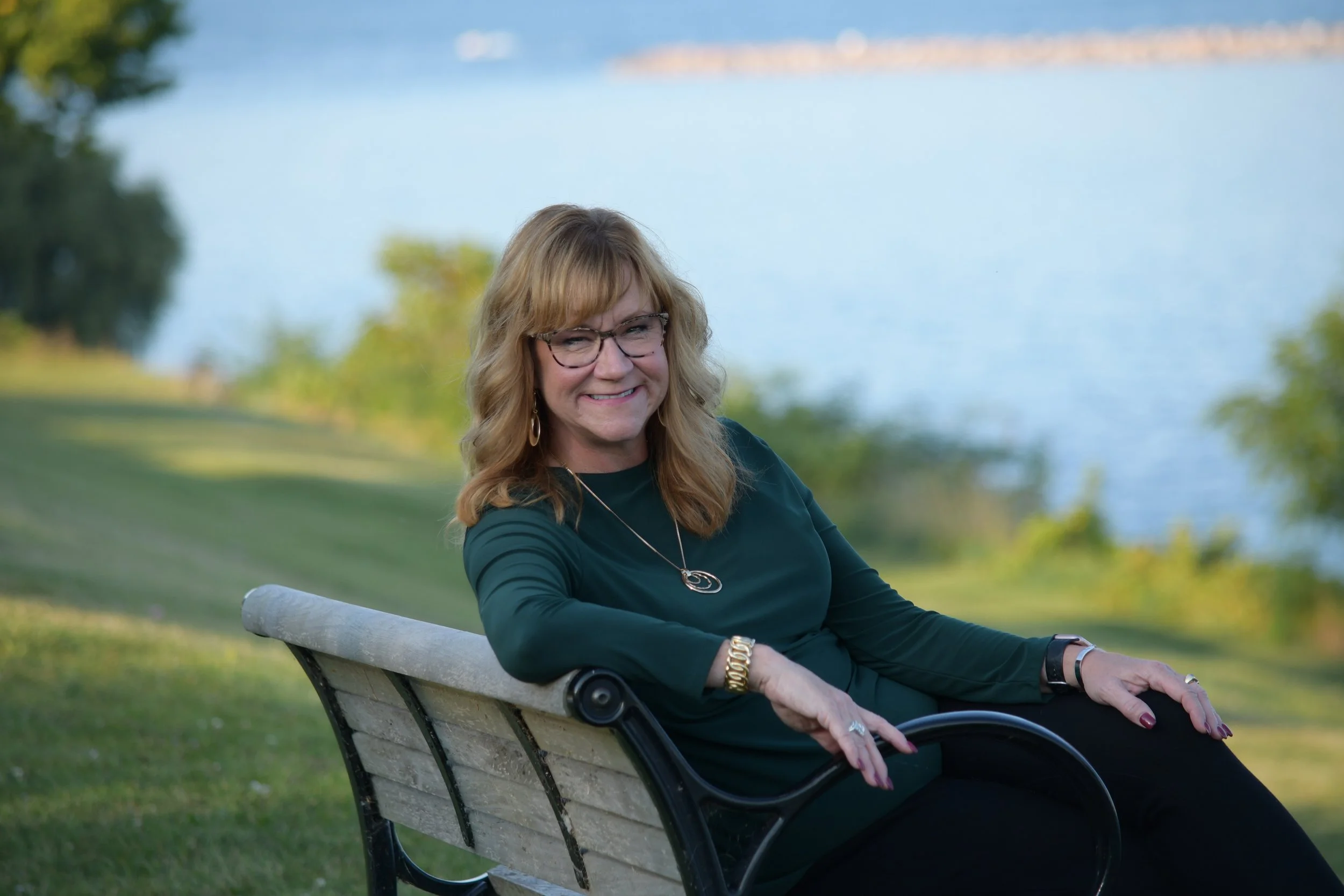 A woman with glasses and blonde hair sitting on a park bench near a lake, smiling at the camera.