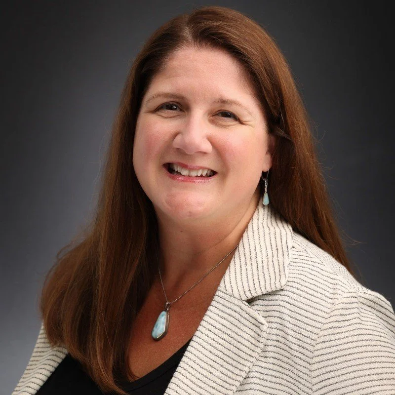 Woman with long brown hair smiling, wearing a white blazer and turquoise jewelry on a dark background.