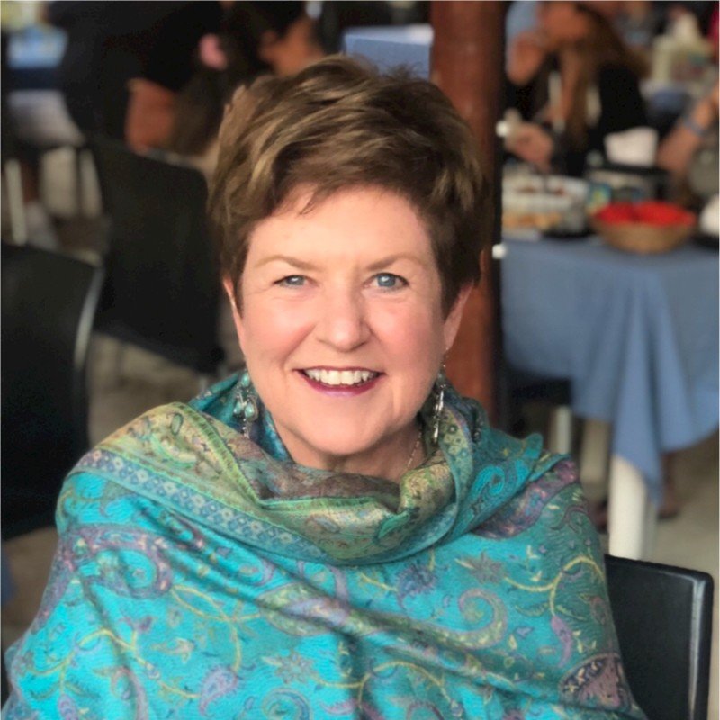 A smiling woman in a colorful, patterned shawl sitting at a table in a busy indoor setting.