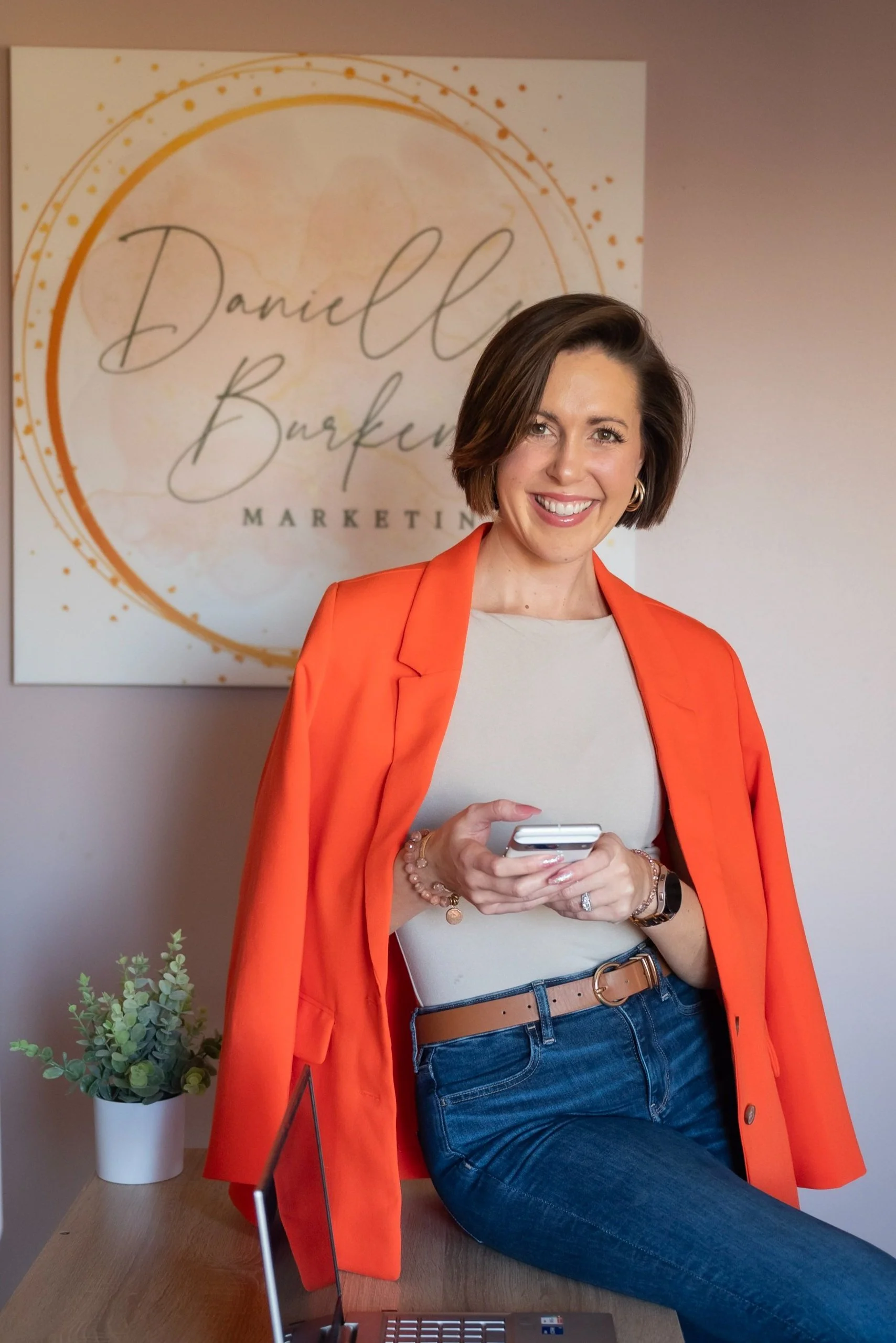 A woman with short brown hair smiling while holding a smartphone, sitting in office space with a sign reading "Danielle Burbank Marketing" in the background, wearing a white top, orange blazer, and blue jeans.