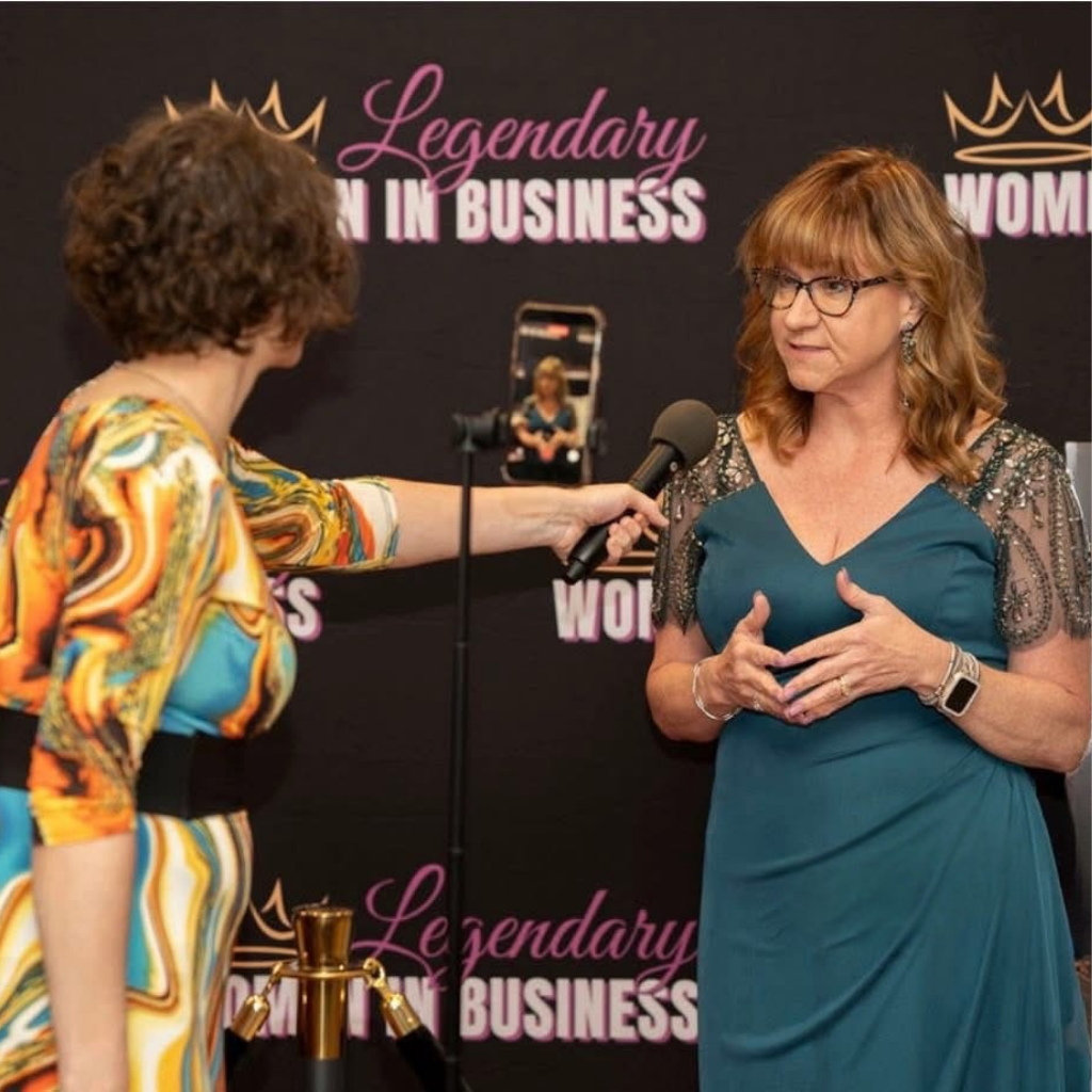 A woman in a teal dress is speaking into a microphone held by another woman during an event. The background displays the words 'Legendary Woman in Business'.