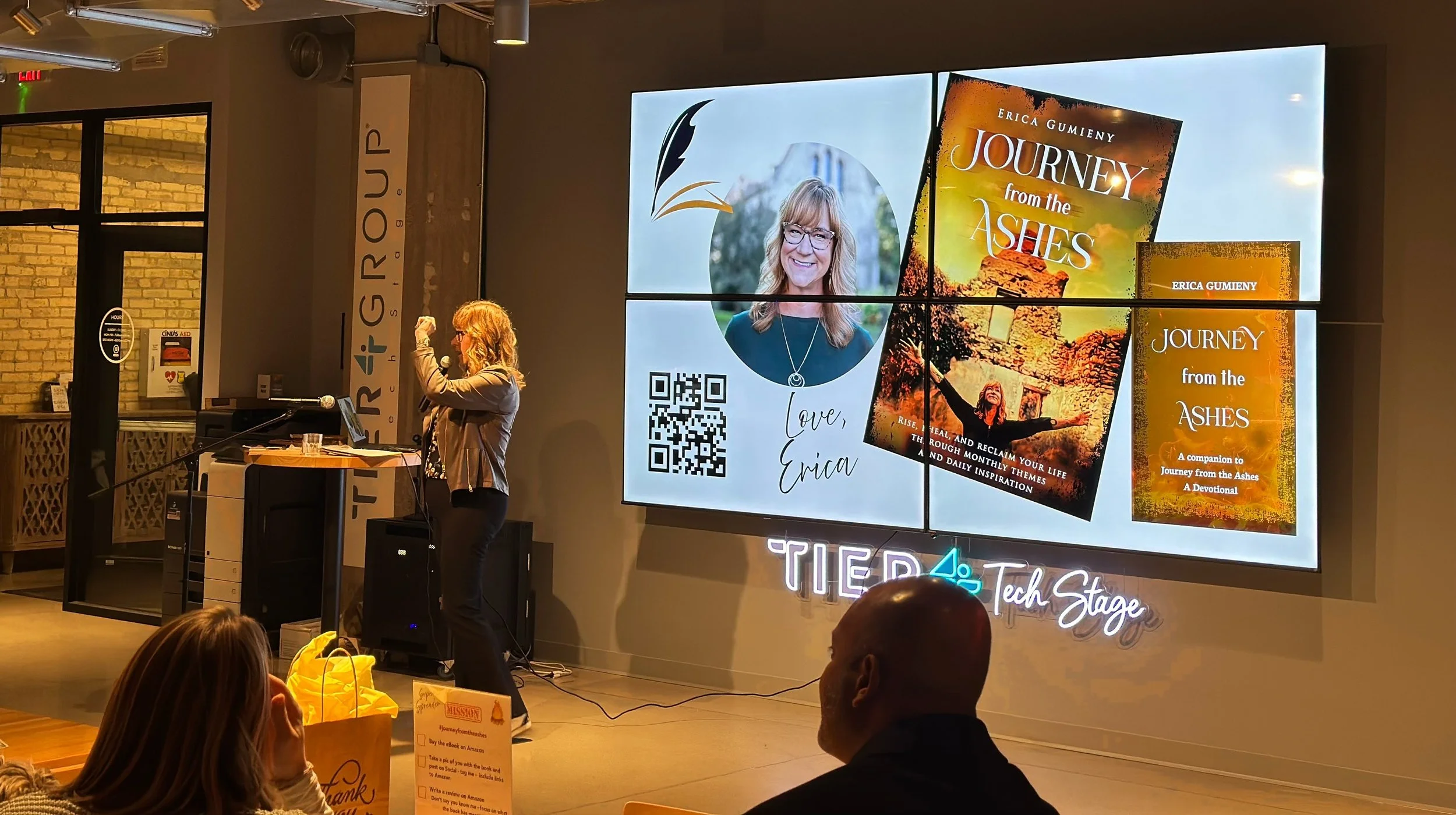 A woman giving a presentation next to a large screen displaying the book covers of "Journey from the Ashes" by Erica Gumieny and other promotional material. The audience is seated in front of her.