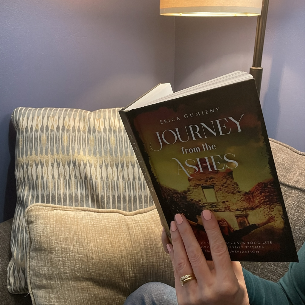 A person reading the book titled "Journey from the Ashes" by Erica Gumieny, sitting on a sofa with patterned and textured pillows, next to a floor lamp in a cozy room with purple walls.