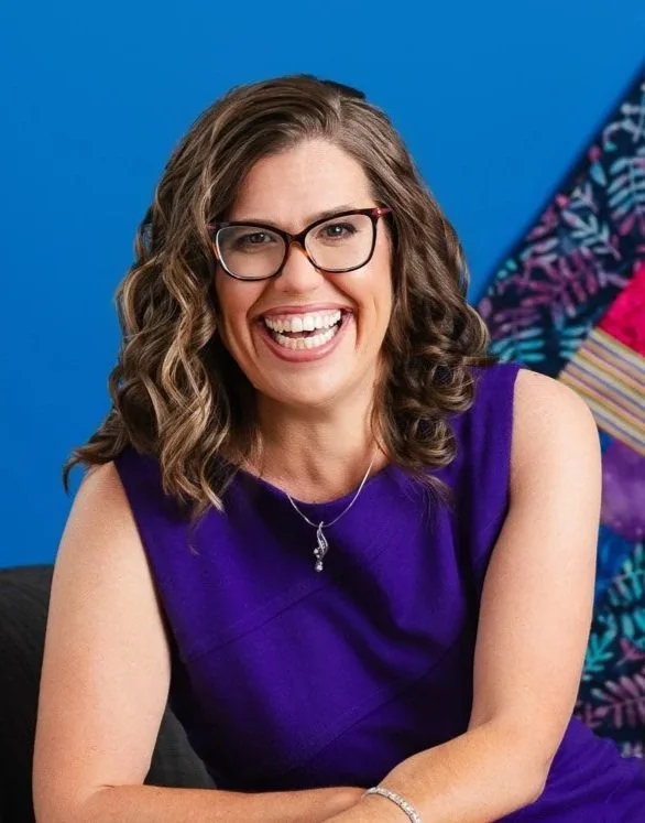 A woman with shoulder-length brown hair, smiling slightly, wearing a black top and a beaded necklace, posing in front of a dark wooden background.