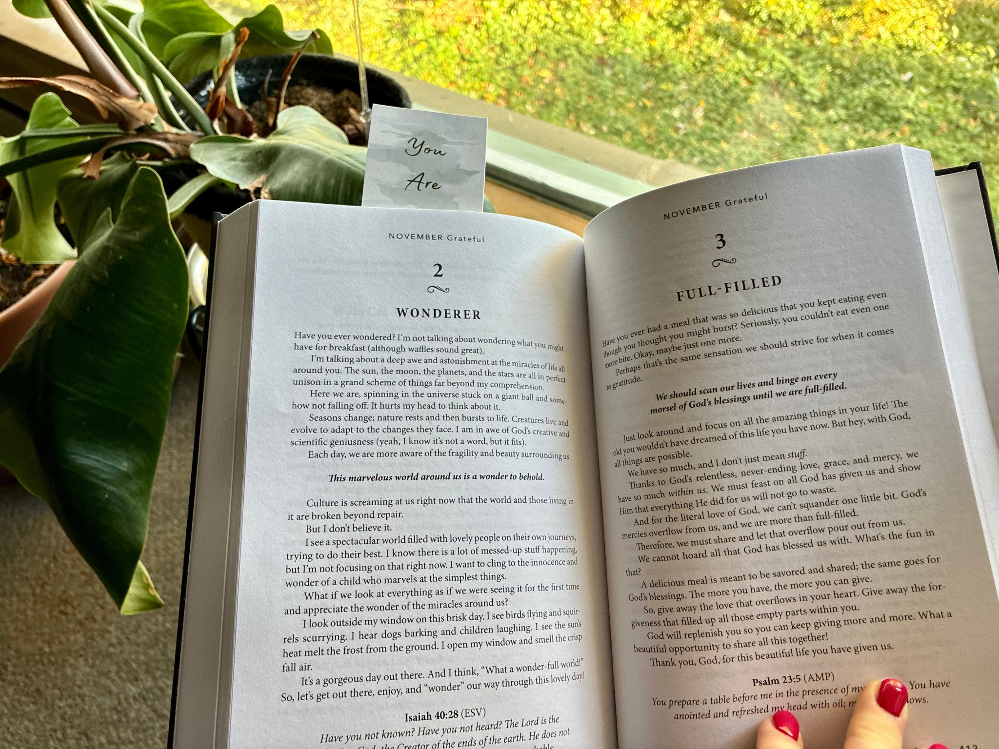 An open book on a window sill with green plants nearby. The book's pages display text about gratitude and wonder, and there is a small note card behind the book that says "You Are". Outside the window, there is a blurred view of green foliage.