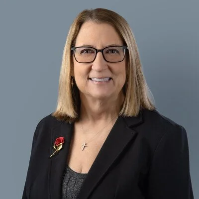 A middle-aged woman with shoulder-length brown hair and glasses, wearing a black blazer with a pin on the lapel, smiling against a plain gray background.