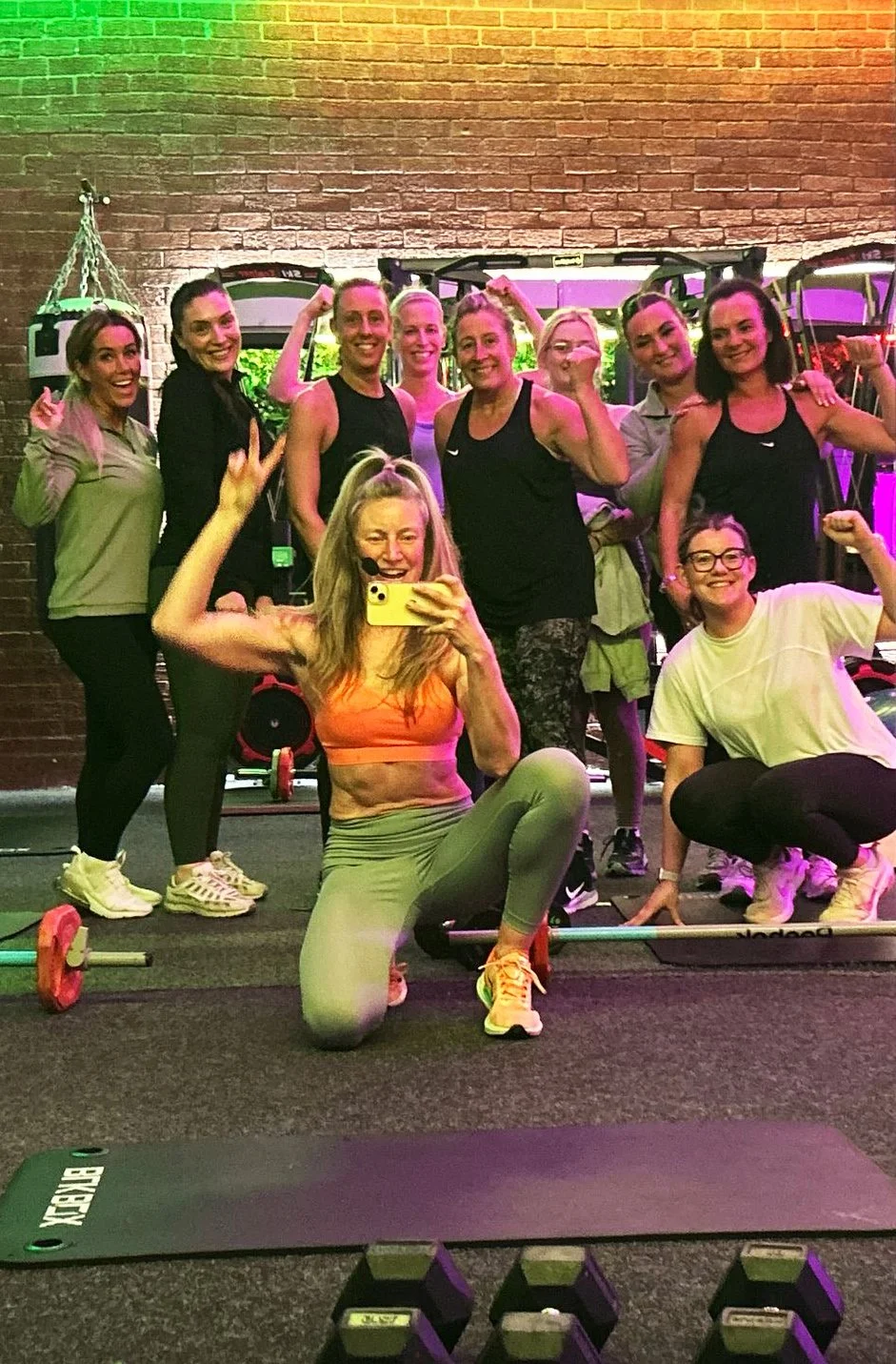 Group of women and a girl posing for a photo in a gym, with fitness equipment and colorful lighting in the background.