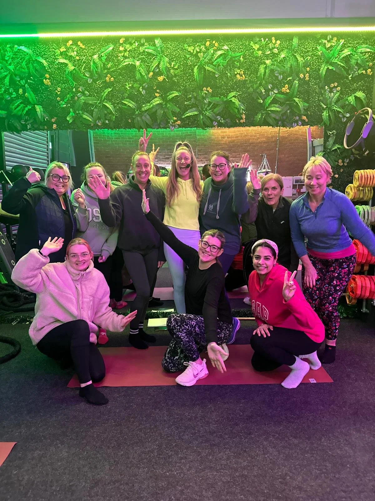 A group of smiling women, some making peace signs or raising their hands, gathered in a fitness studio with colorful lighting, fitness equipment, and a wall of plants in the background.