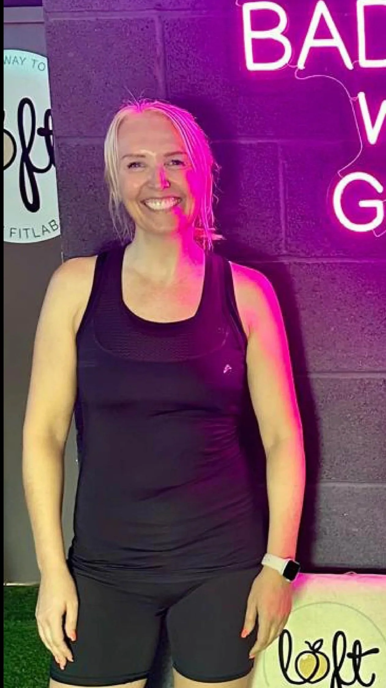 A woman smiling in front of a neon sign that reads 'BAD W' above her head, dressed in black athletic wear, wearing a watch, with long blonde hair, against a dark background.