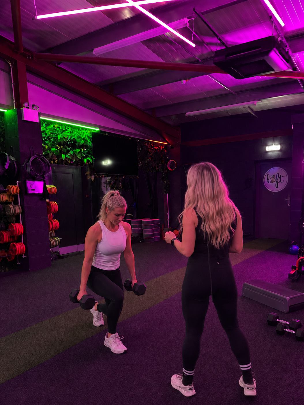 Two women exercising with dumbbells in a gym with purple and pink neon lighting.