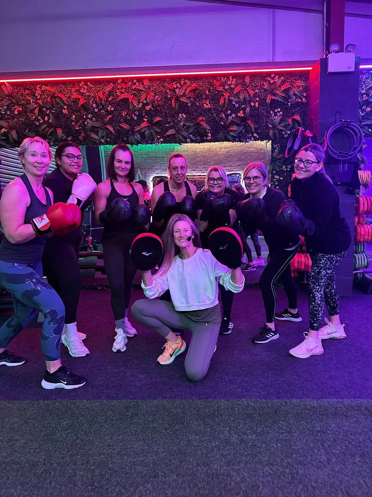 Group of women in a fitness class, posing with boxing gloves and exercise equipment in a gym with colorful lighting.
